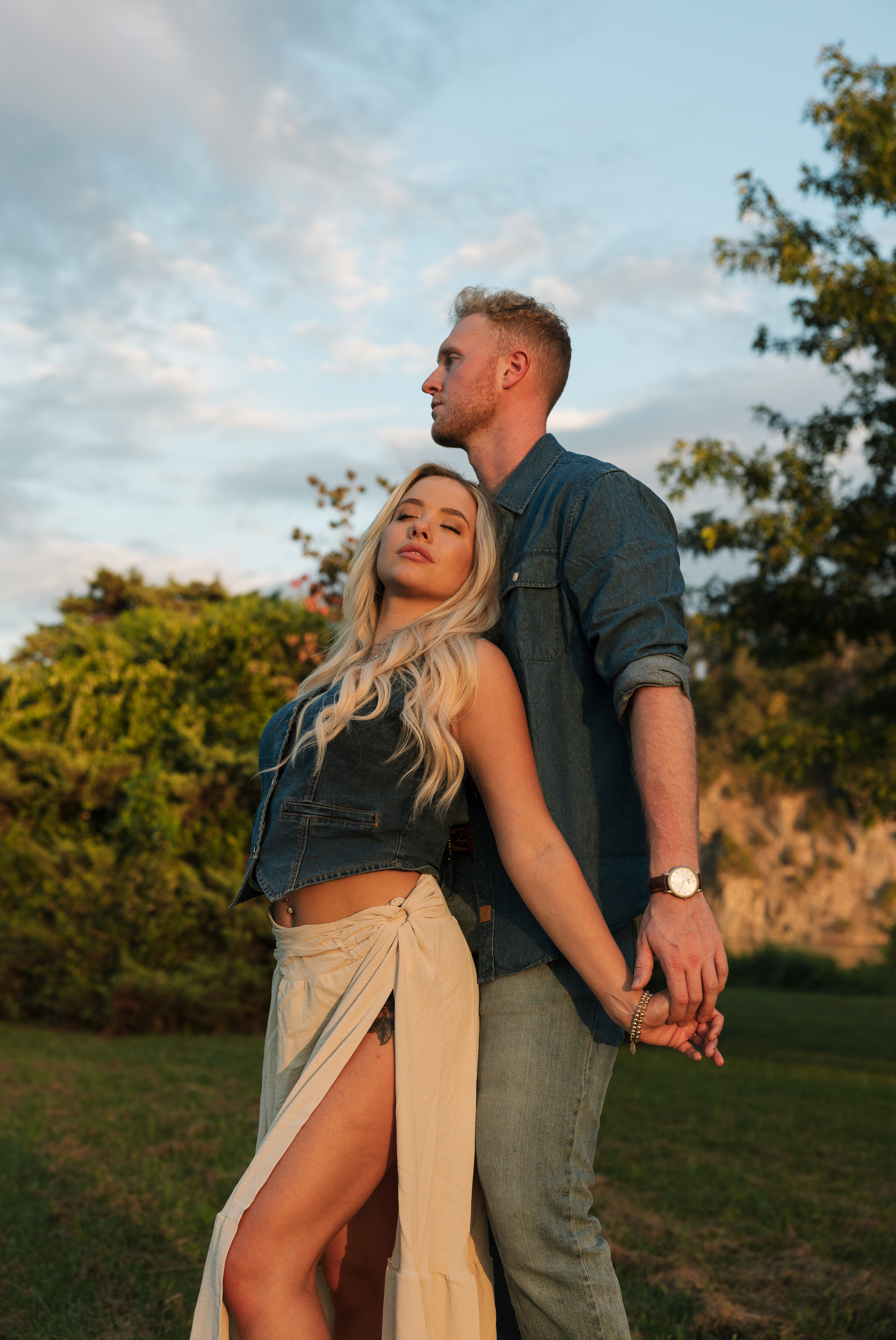 Engaged couple holding hands and posing in a sunlit open field during a modern Western engagement session in Knoxville, with bold, dramatic lighting and stylish cowboy boots