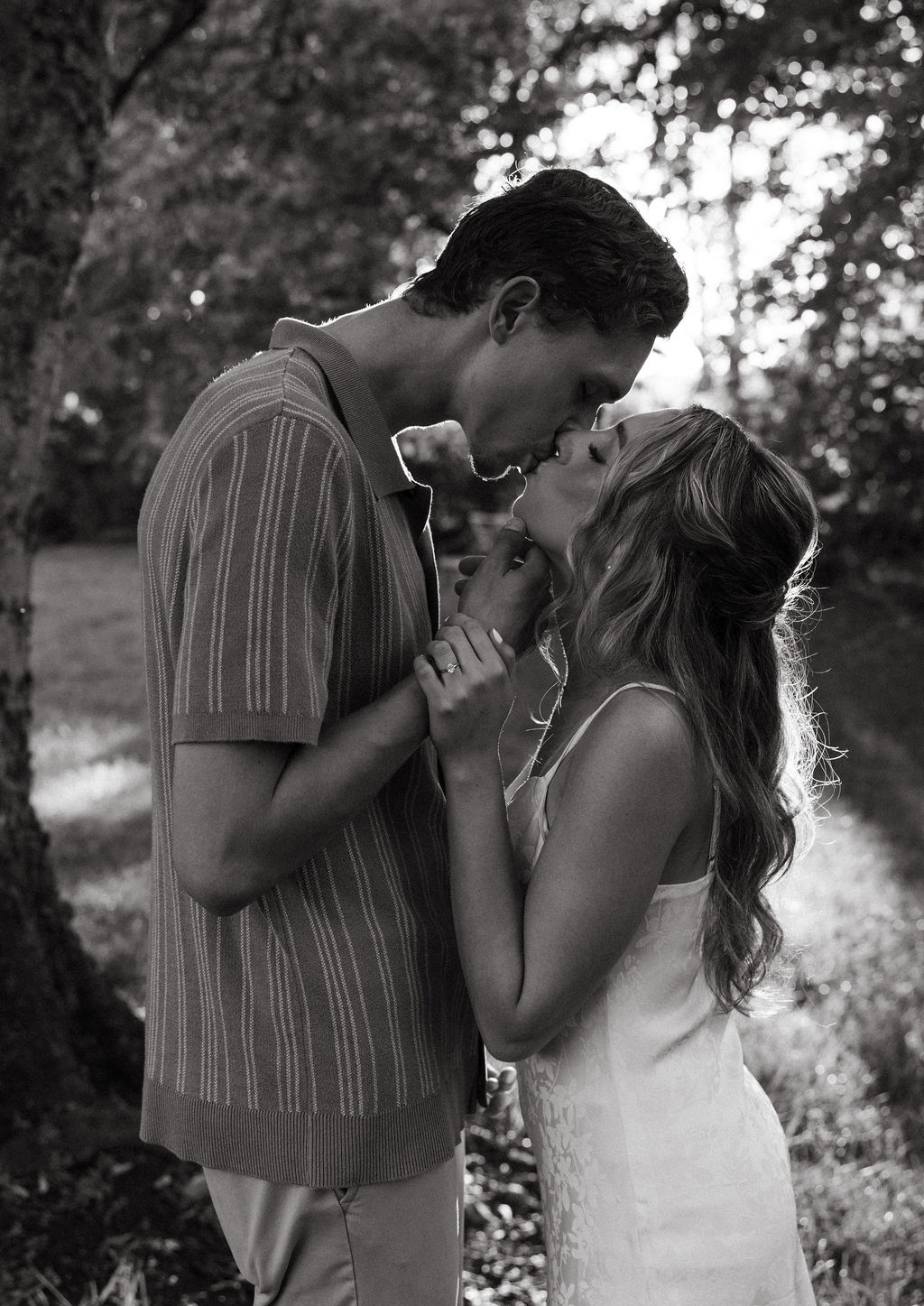 A close-up black and white image of a couple kissing tenderly in a wooded area with soft sunlight in the background, captured during engagement photos by a Knoxville photographer.