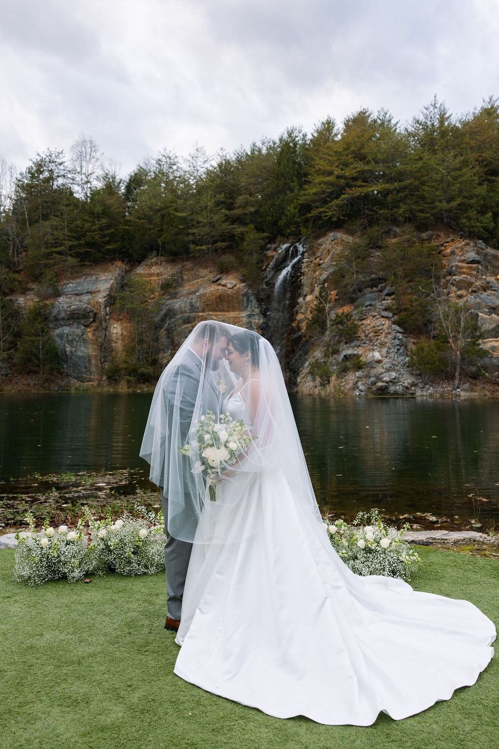 Bride and groom stand beneath a veil, embracing in front of a serene lake and rocky waterfall backdrop, part of a romantic scene curated by a Knoxville wedding planner.