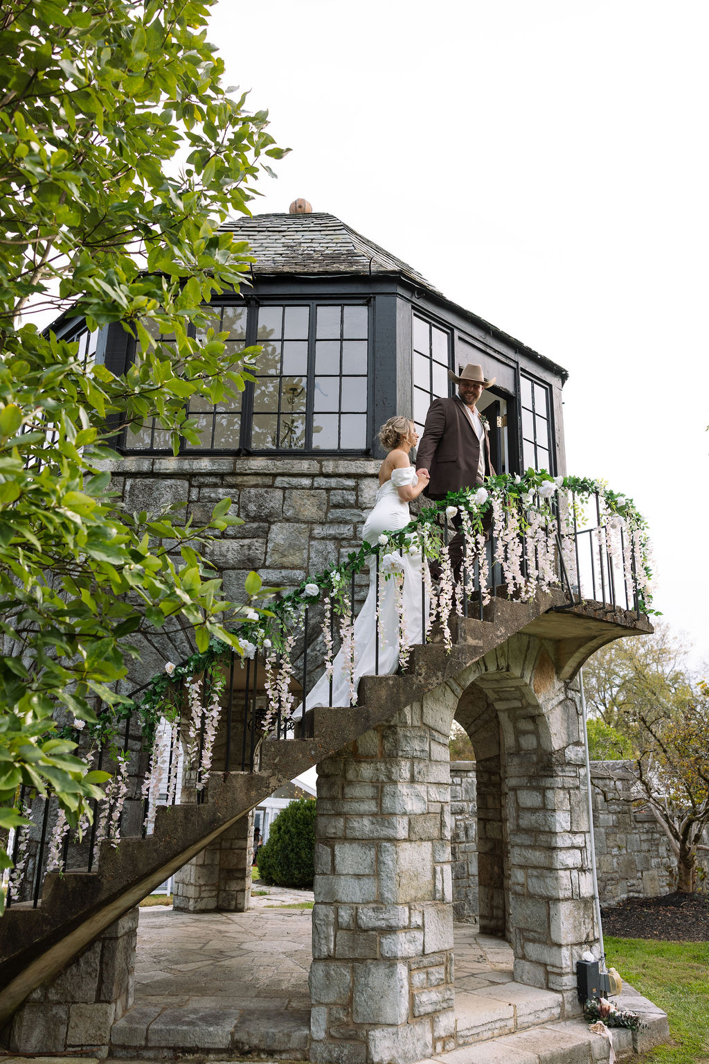The bride and groom descend the winding stone staircase of The Kincaid House tower, hand in hand. The railing is adorned with cascading white florals and greenery, echoing the romantic charm of the venue. The bride’s elegant train trails behind her, while the groom leads with a warm smile under his wide-brimmed hat. The moment feels timeless and cinematic.