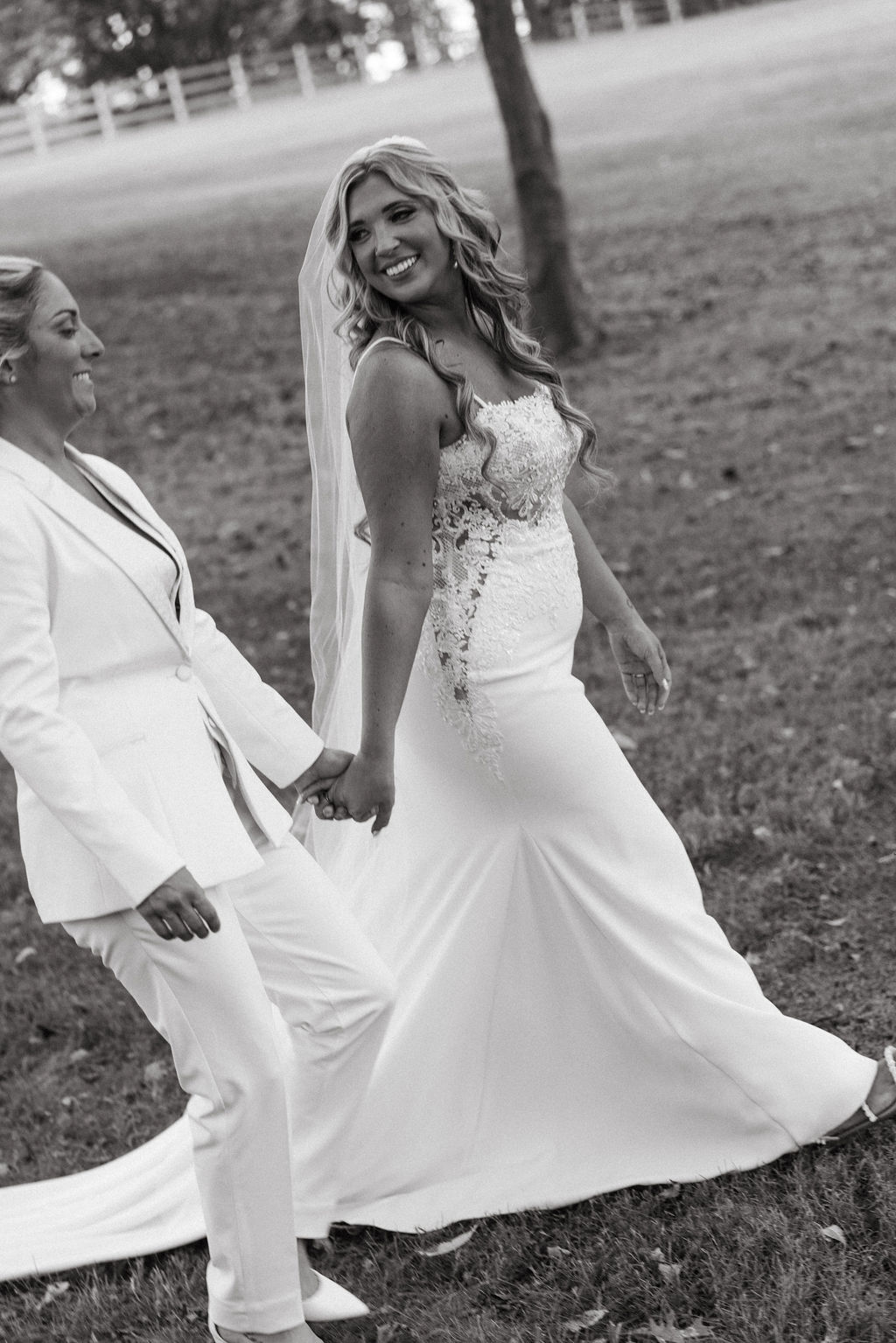 A black and white image of two brides walking hand in hand across the lawn at Marble Gate Farm, both smiling joyfully, captured by a Knoxville wedding photographer.