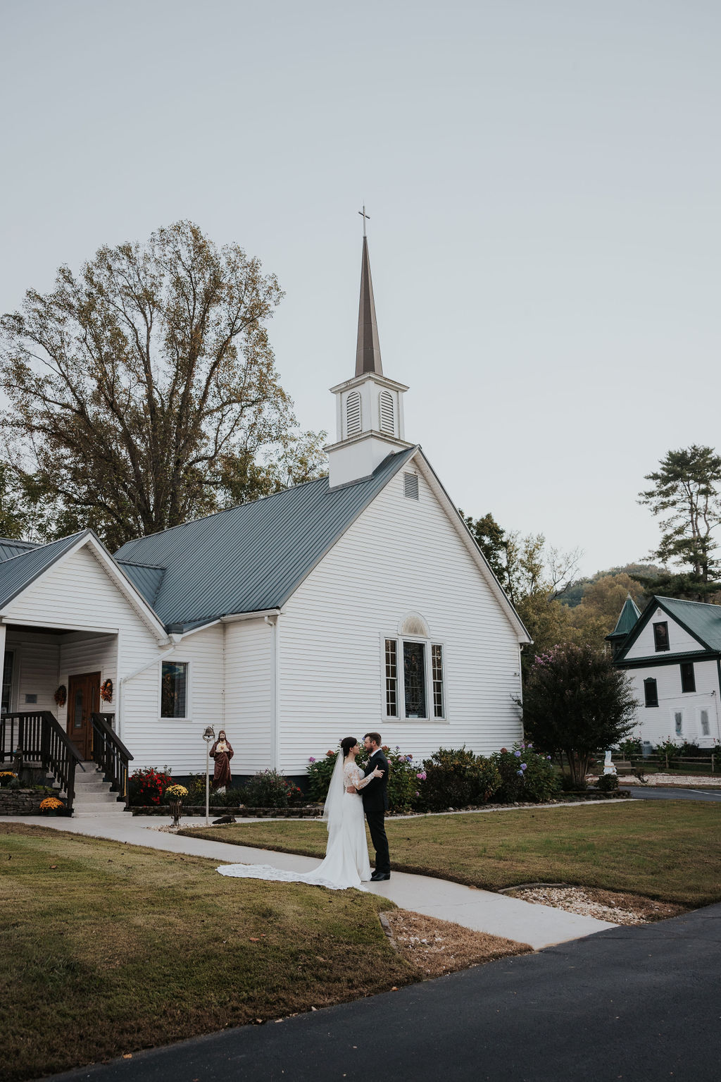 Wide-angle view of a couple embracing in front of a traditional white chapel wedding venue with a tall steeple, surrounded by blooming flowers and mature trees; photographed by a Knoxville wedding photographer