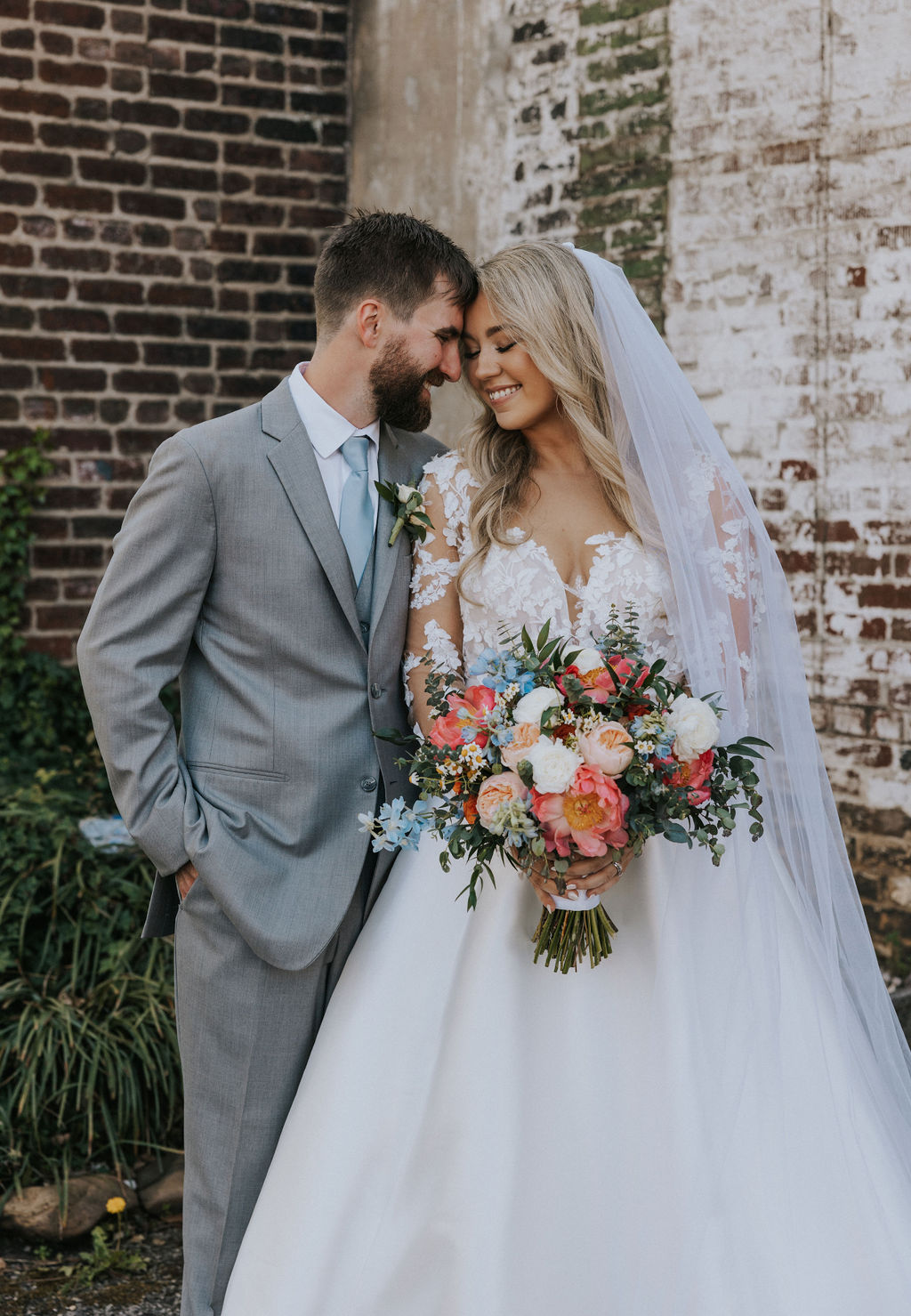 Bride and groom standing close with foreheads touching, smiling as the bride holds a colorful bouquet in front of a textured brick wall at The Press Room Knoxville wedding, photographed by a Knoxville wedding photographer.
