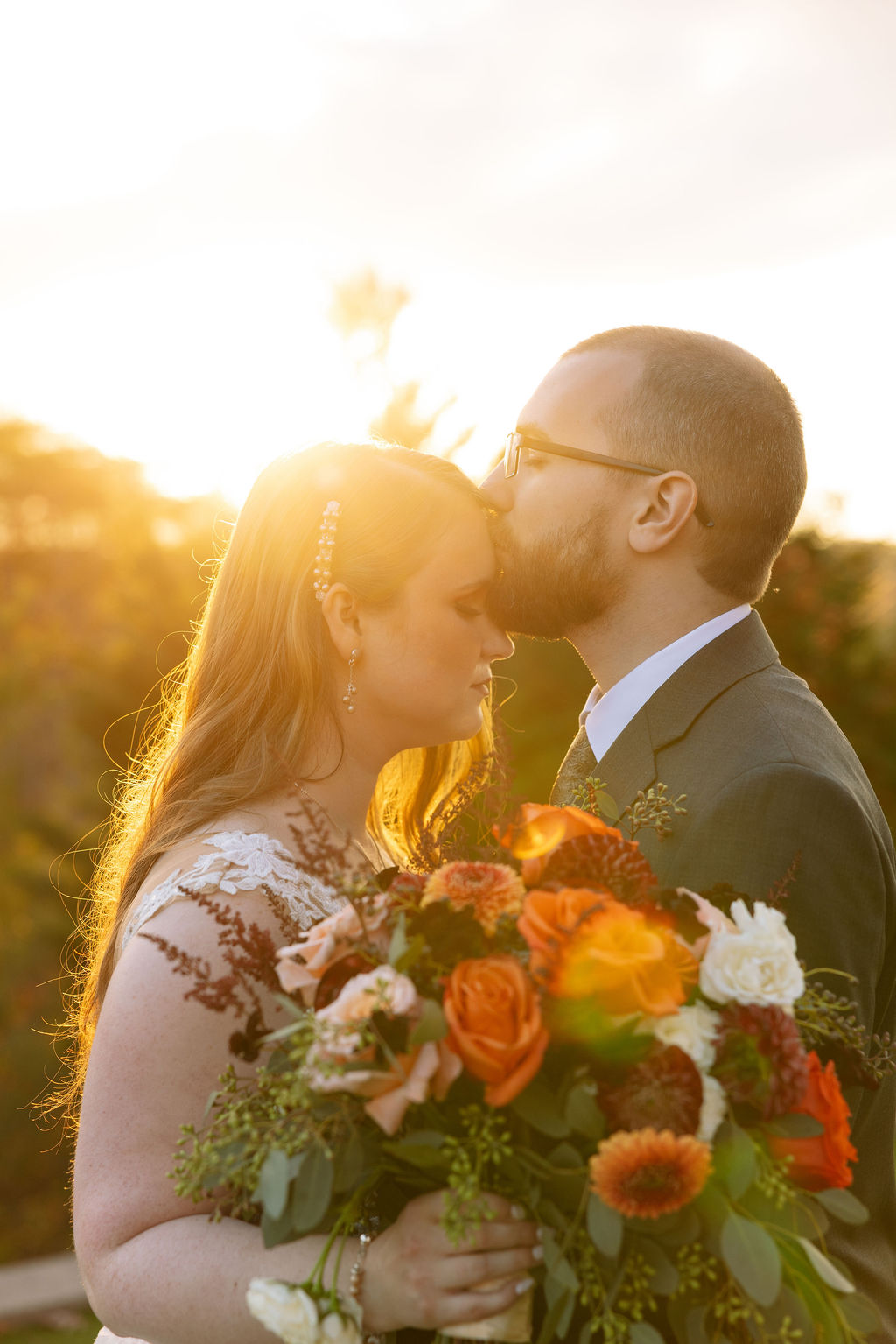 Close-up of the groom kissing the bride’s forehead at sunset, with golden light illuminating her face and vibrant fall bouquet; captured by a Knoxville wedding photographer
