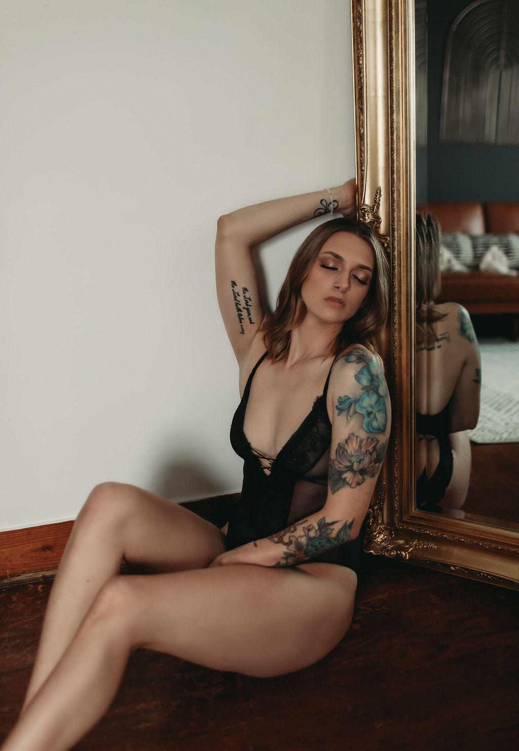 Boudoir photography portrait of woman sitting beside vintage gold mirror in black bodysuit with soft natural light.