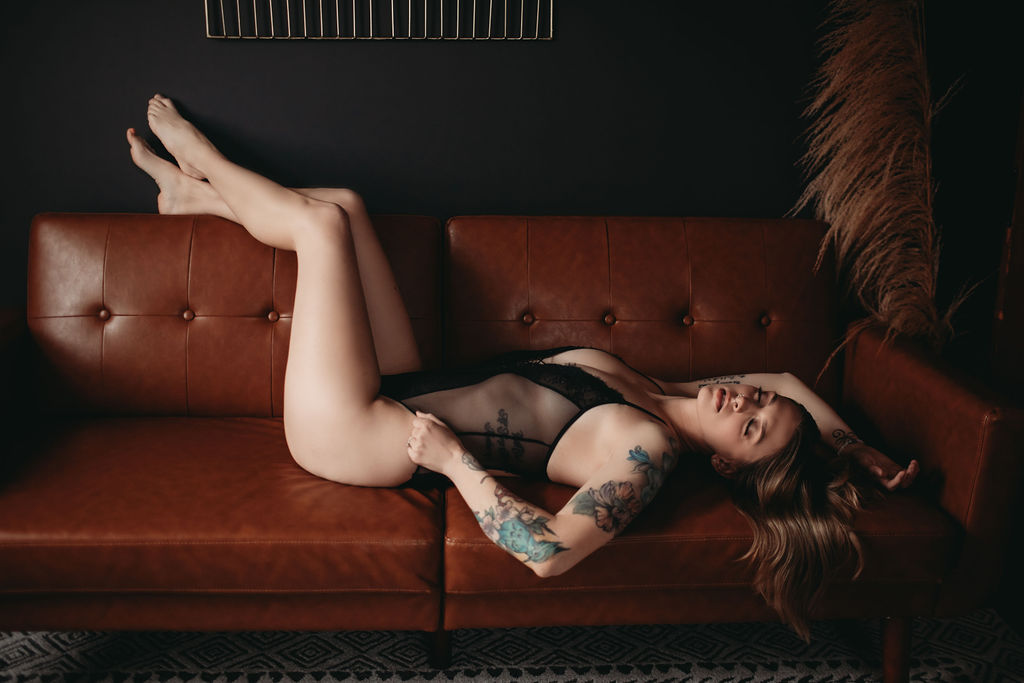 Woman wearing black bodysuit with eyes closed while laying on brown leather couch with legs in the air during boudoir photography shoot. 