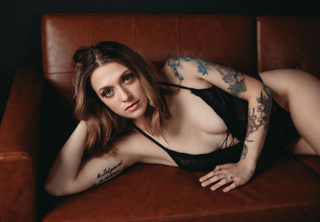 Intimate portrait of woman in black lace bodysuit lying on brown leather couch with soft, moody lighting.