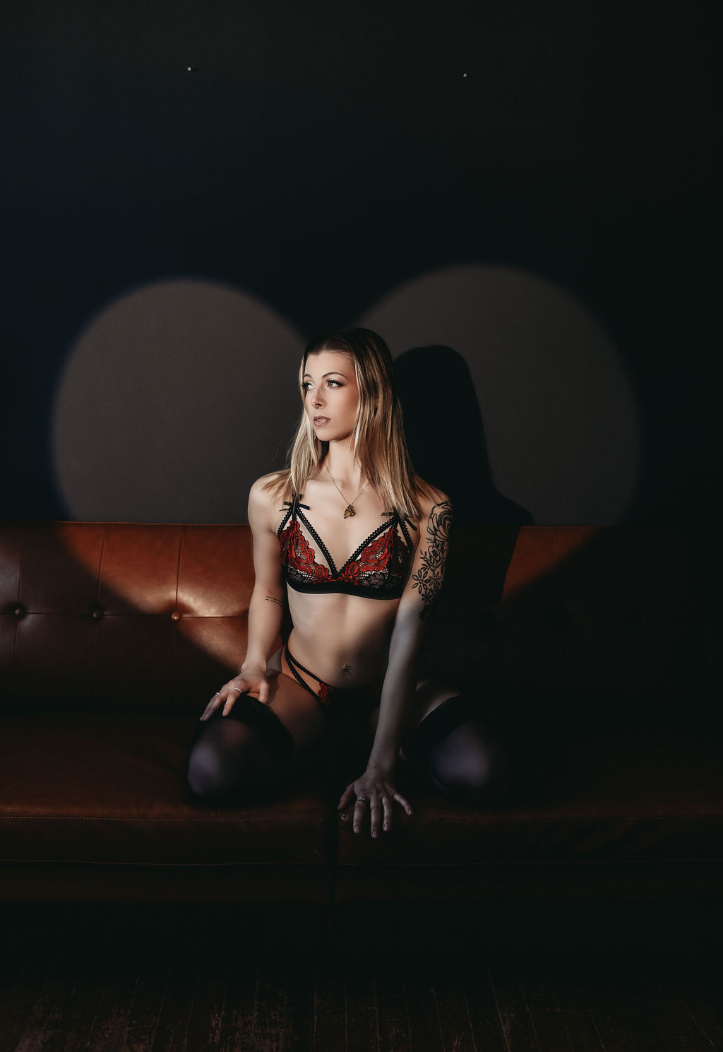 Woman seated on a leather couch in red and black lingerie, photographed by a boudoir photographer with circular shadow patterns behind her.