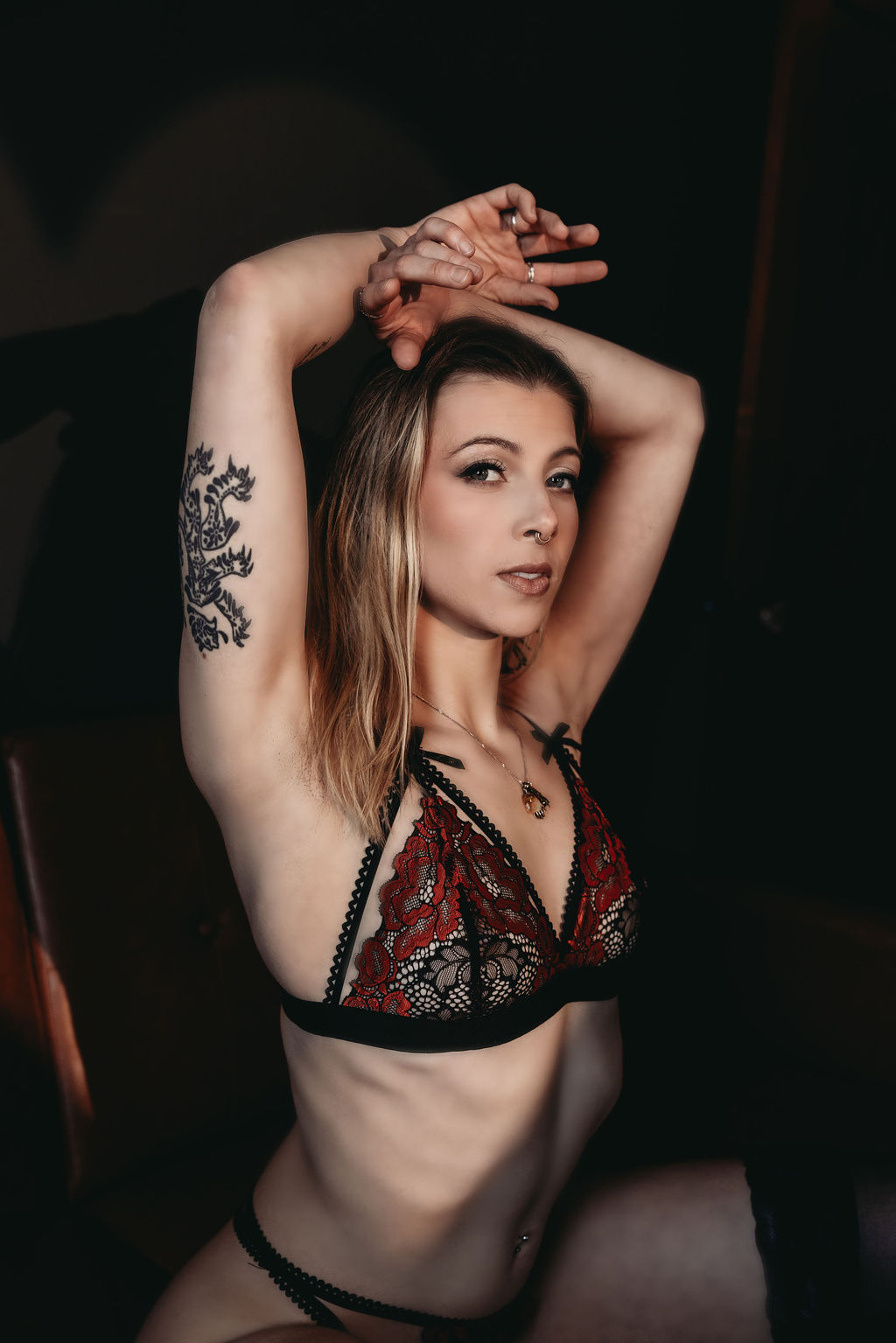 Close-up portrait of a woman in red lace lingerie with arms raised above her head, softly lit against a dark studio backdrop.