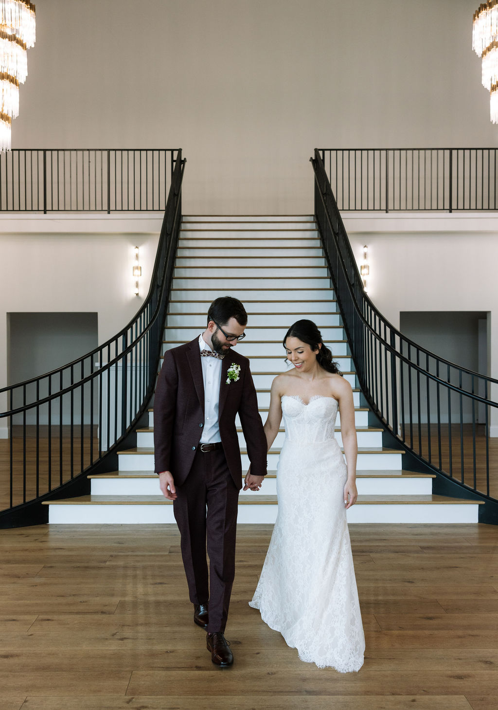 Bride and groom walking hand in hand in front of the grand staircase at Nichols Heir, calm indoor portrait that shows what do wedding planners do to maintain flow on a rain-filled wedding day.