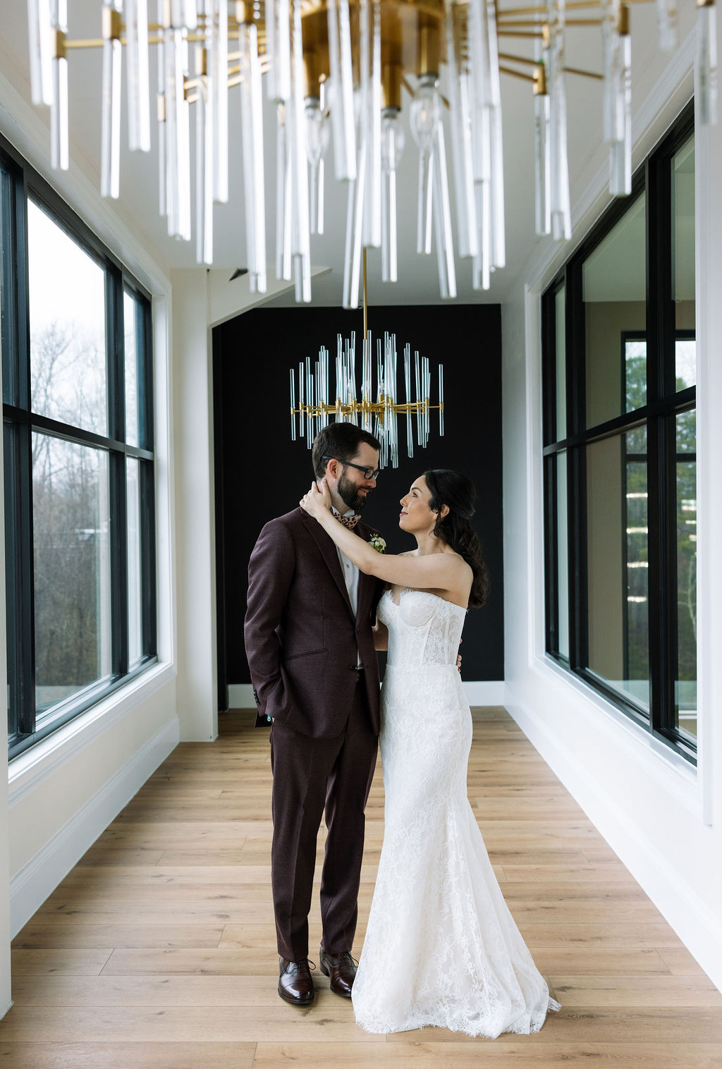 Bride and groom standing beneath modern chandeliers in the Glass House at Nichols Heir, true-to-color indoor wedding portraits during a rain-adjusted timeline.