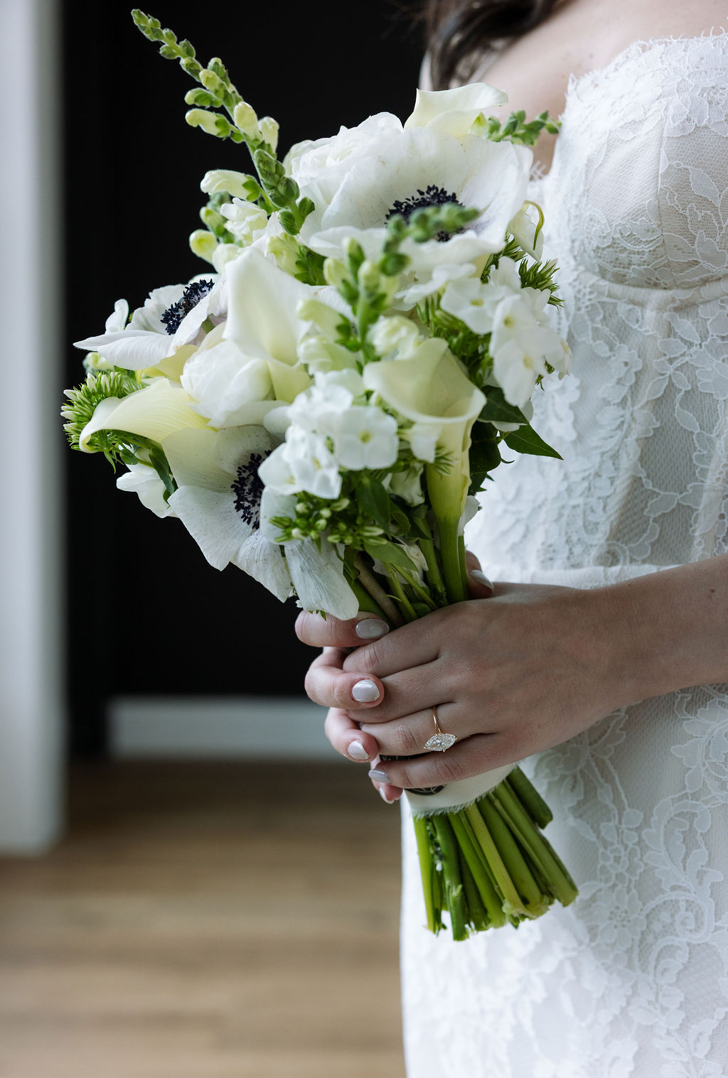 Close-up of bride holding a white bouquet of anemones and roses, lace wedding gown and engagement ring visible in a softly lit indoor portrait.