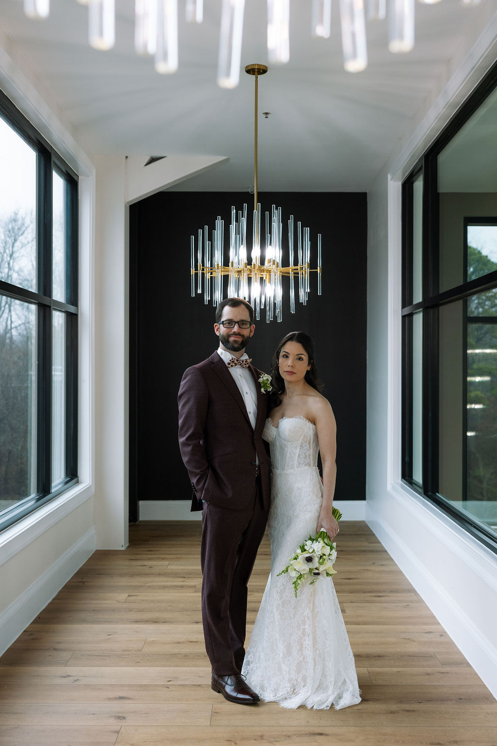 Bride and groom standing together under a chandelier in the glass house as newly married husband and wife.