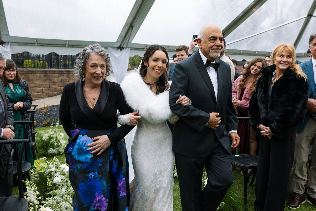 Bride walking down the aisle between her parents under a rain-covered tent, a steady ceremony moment that shows what do wedding planners do to coordinate timing and guest flow.