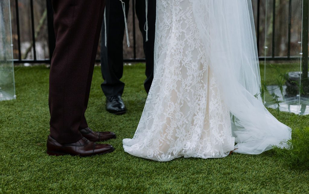 Close-up of lace wedding dress hem and groom’s shoes on wet grass during a rainy Tennessee ceremony setup.