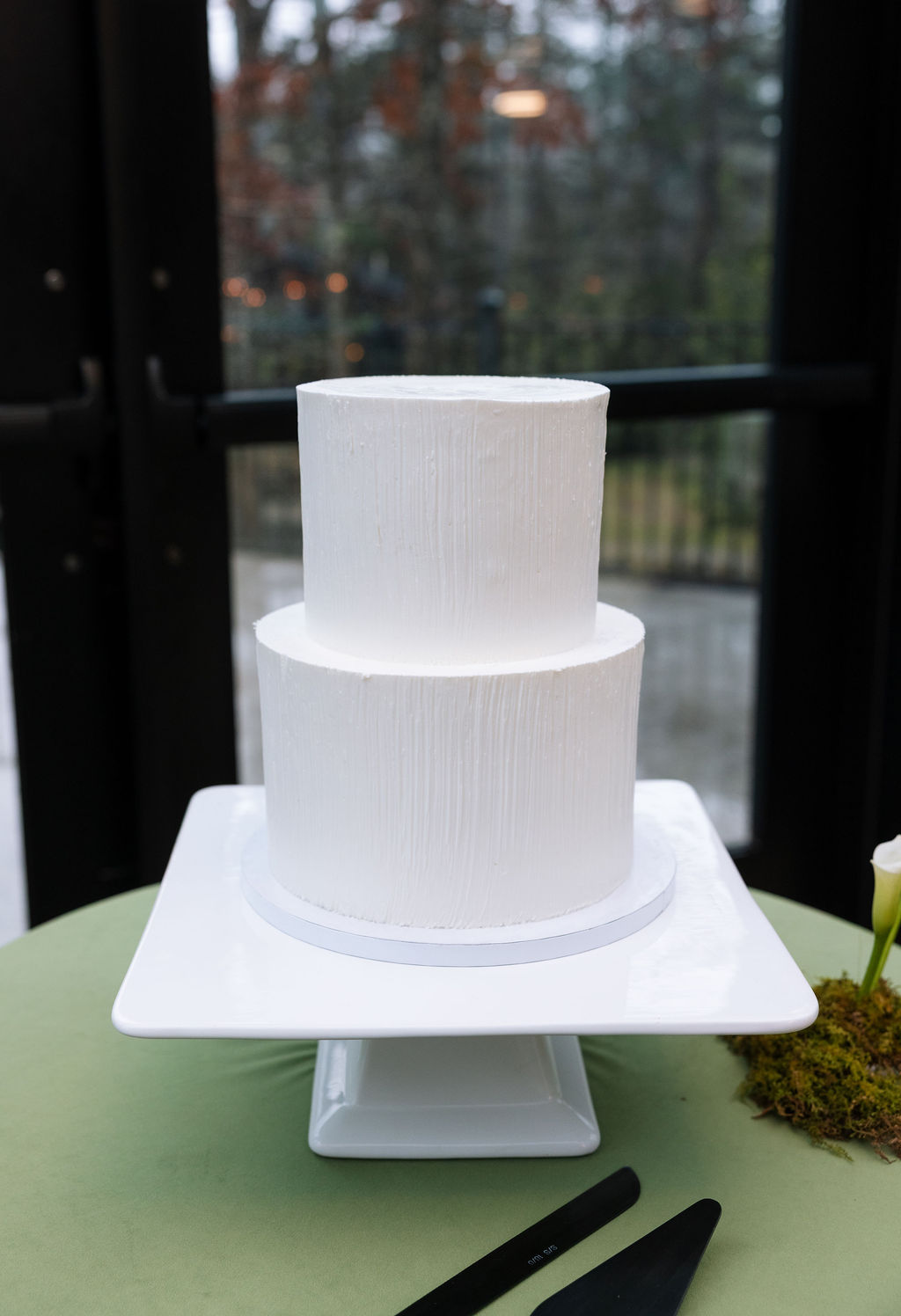 Minimal two-tier white wedding cake displayed on a pedestal in front of floor-to-ceiling windows at a Tennessee indoor reception.