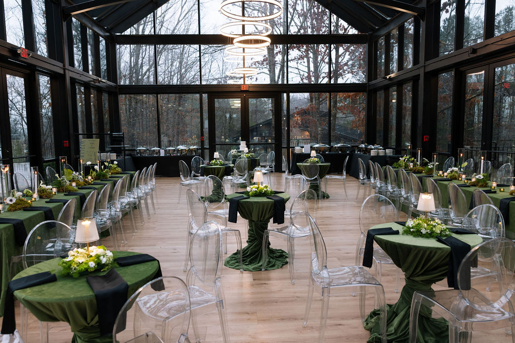 Wide view of the Glass House reception space with green linens, clear chairs, and candlelit tables, demonstrating what do wedding planners do to manage layout and atmosphere.
