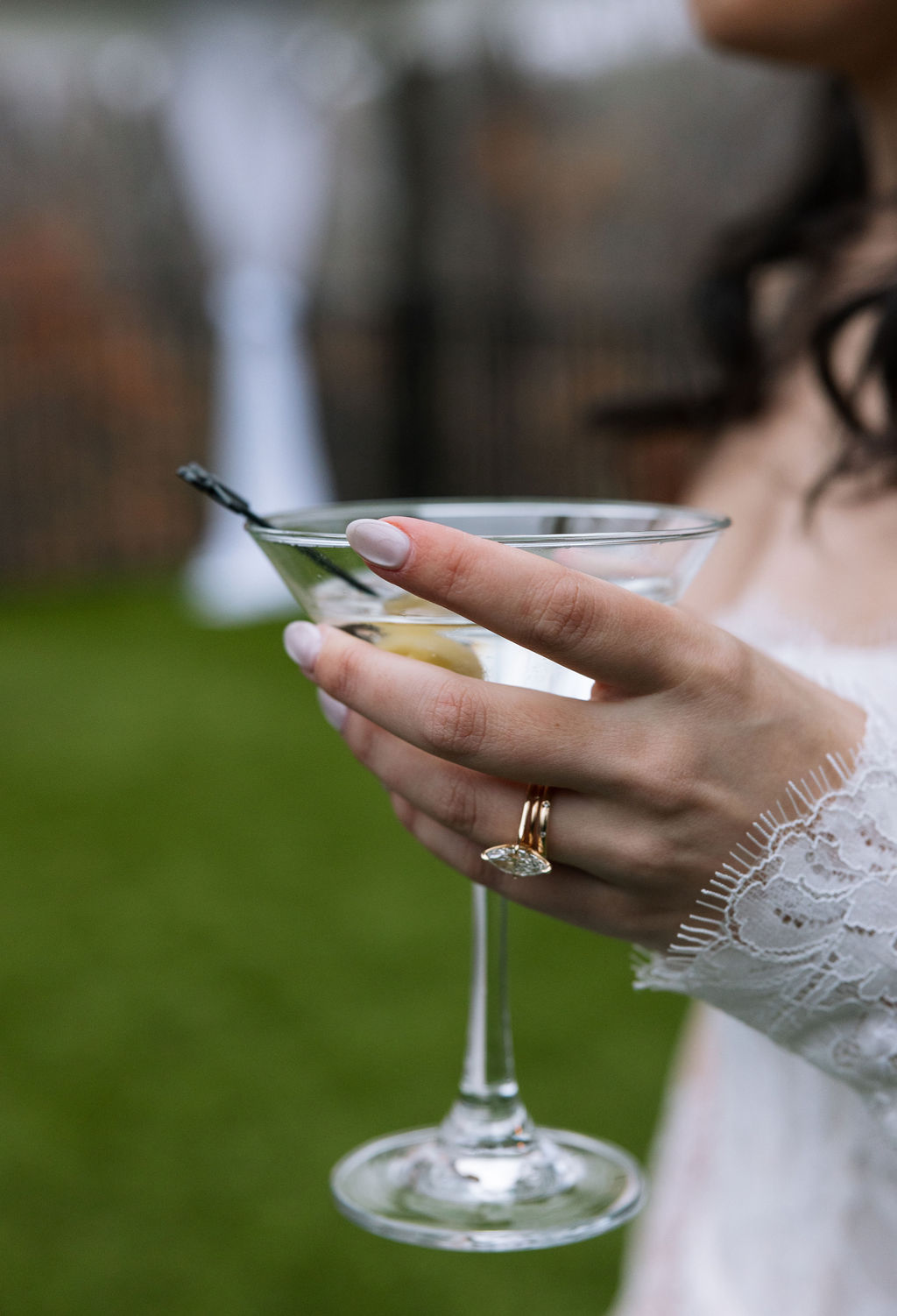 Bride holding a martini glass outdoors after the ceremony, lace sleeve and engagement ring visible in a relaxed cocktail hour portrait.