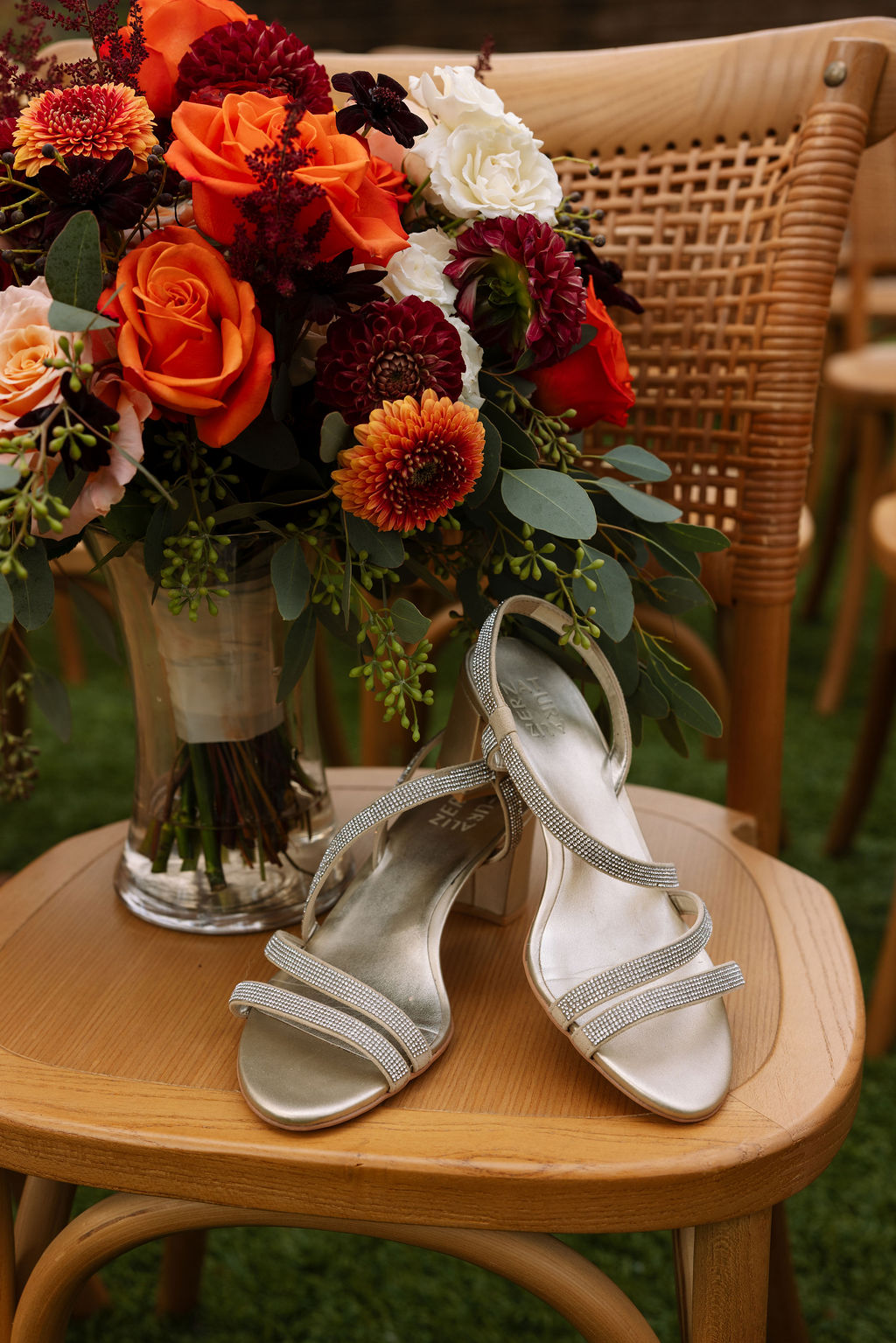 Fall colored bridal bouquet in vase of water sitting beside bride's wedding shoes on rustic chair.