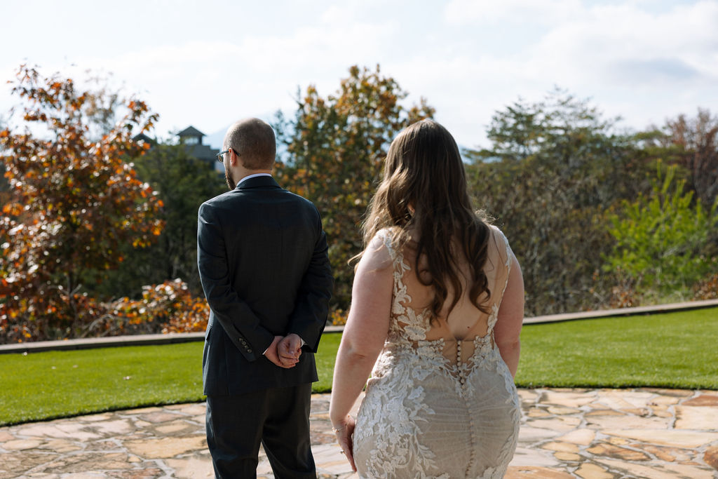 Bride approaching the groom from behind during their first look, with fall trees and open lawn ahead.