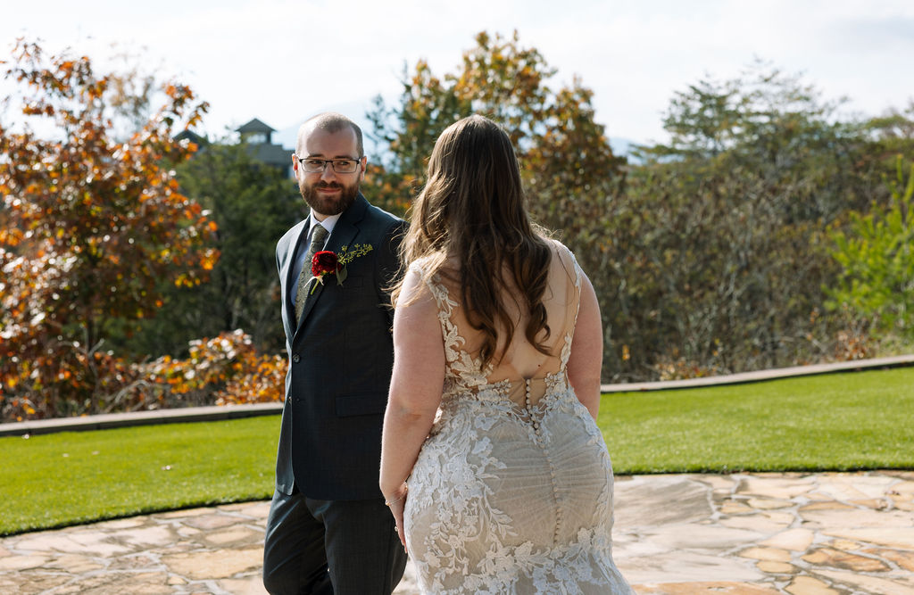Groom turning to see the bride during their first look, captured on a stone patio with fall foliage beyond.
