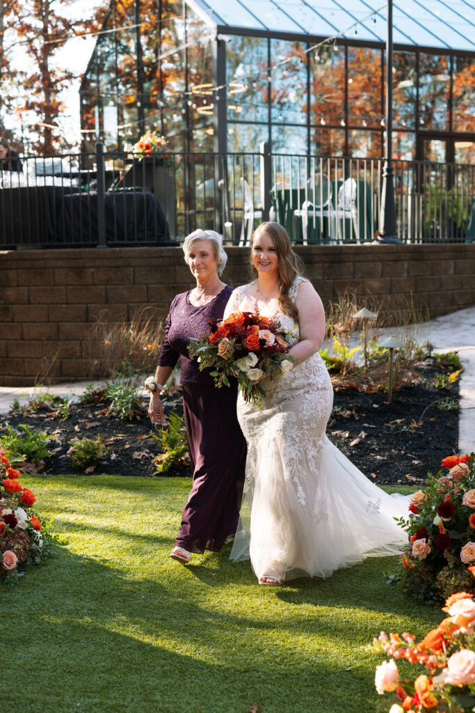 Bride walking arm in arm with her mother toward the ceremony, holding a fall bouquet and smiling.