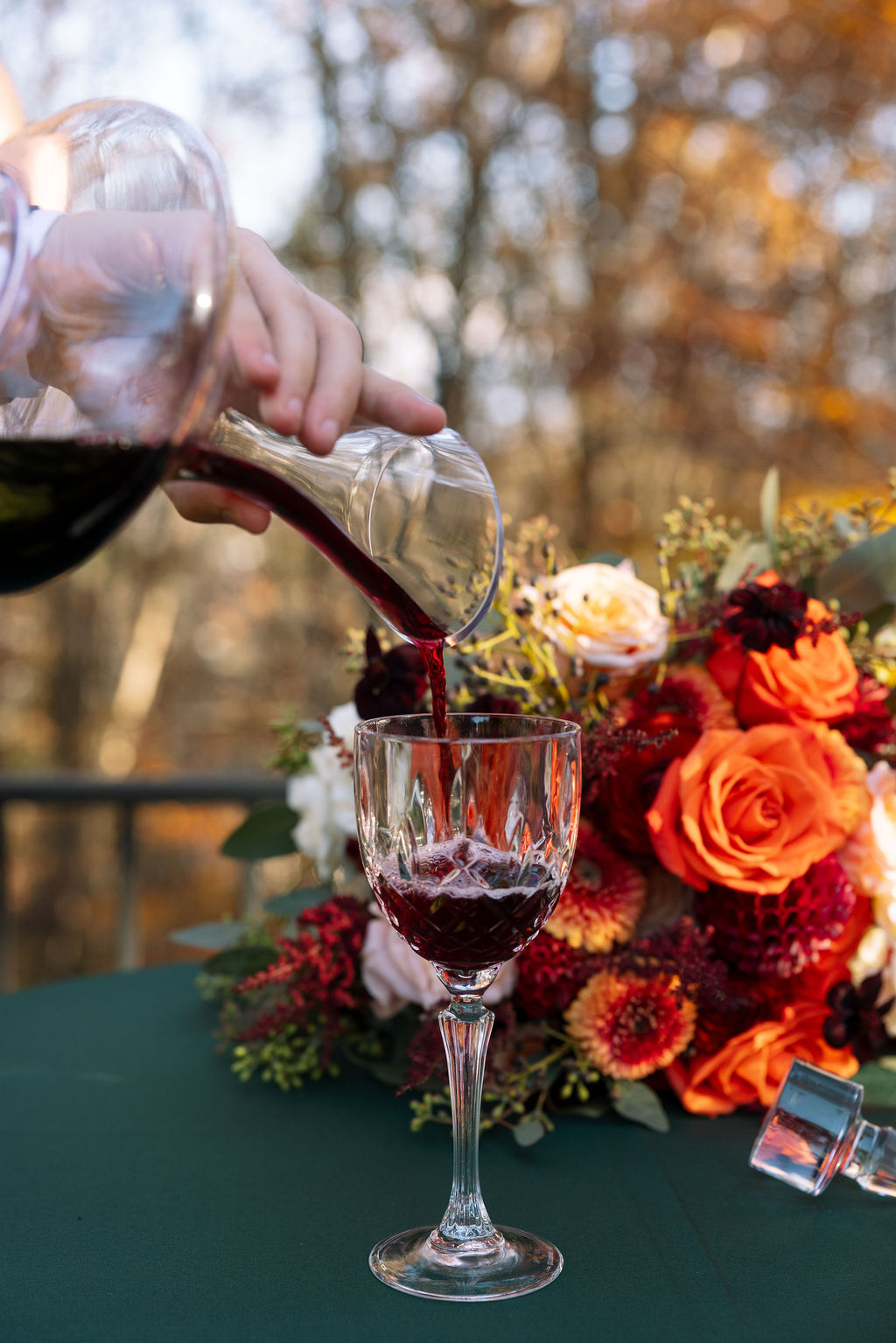 Red wine being poured into a glass beside an autumn floral arrangement during an outdoor wedding celebration.