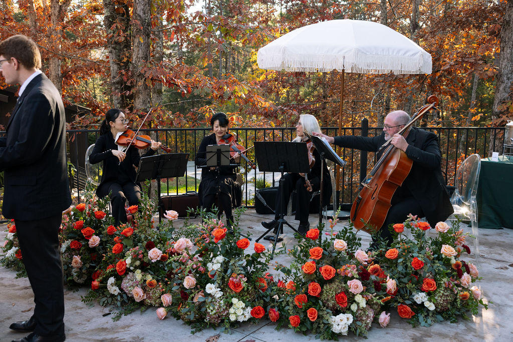 Live string musicians performing during an outdoor fall wedding ceremony, surrounded by florals and autumn color.