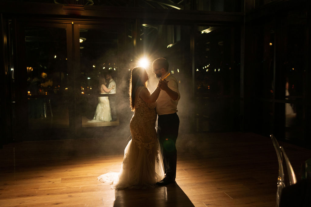 Bride and groom sharing their first dance on a dimly lit floor, backlit for a dramatic and intimate moment.