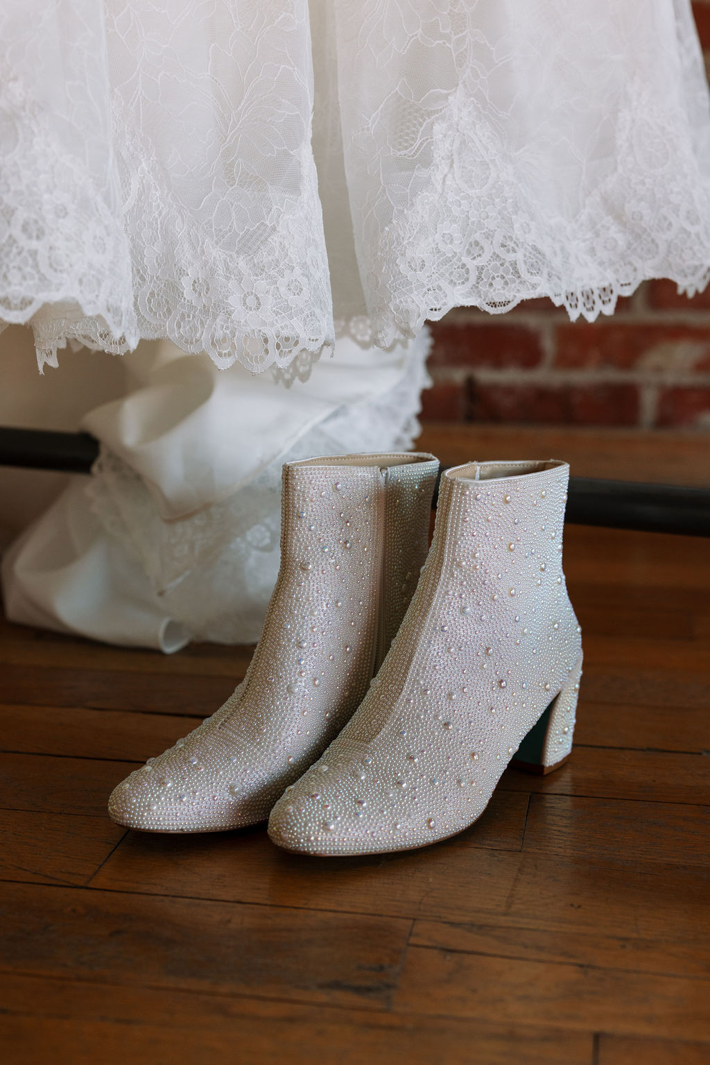 Pearl-studded bridal boots placed beneath a lace wedding dress against exposed brick wall