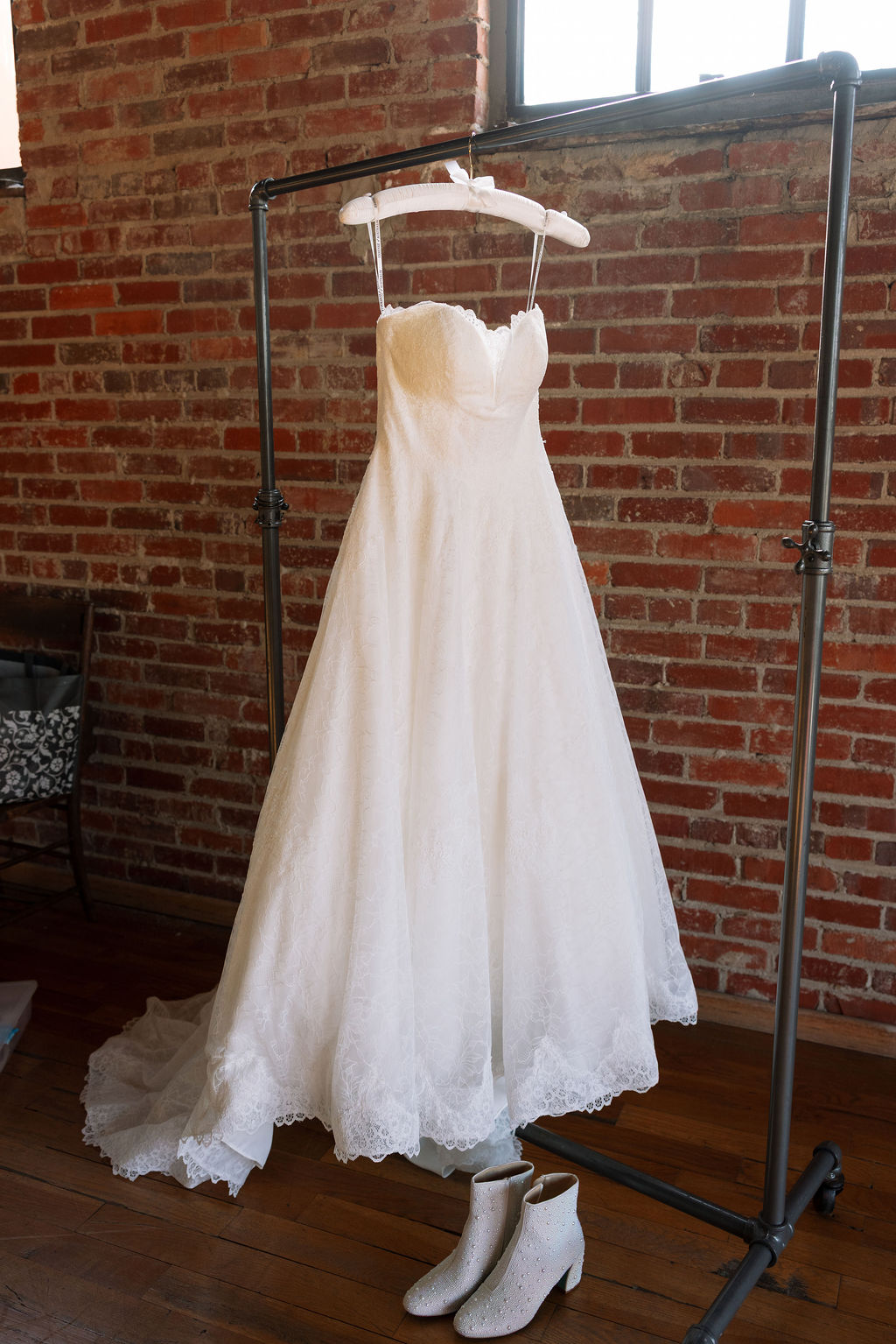 Lace wedding dress hanging on a rack with bridal boots below against exposed brick wall