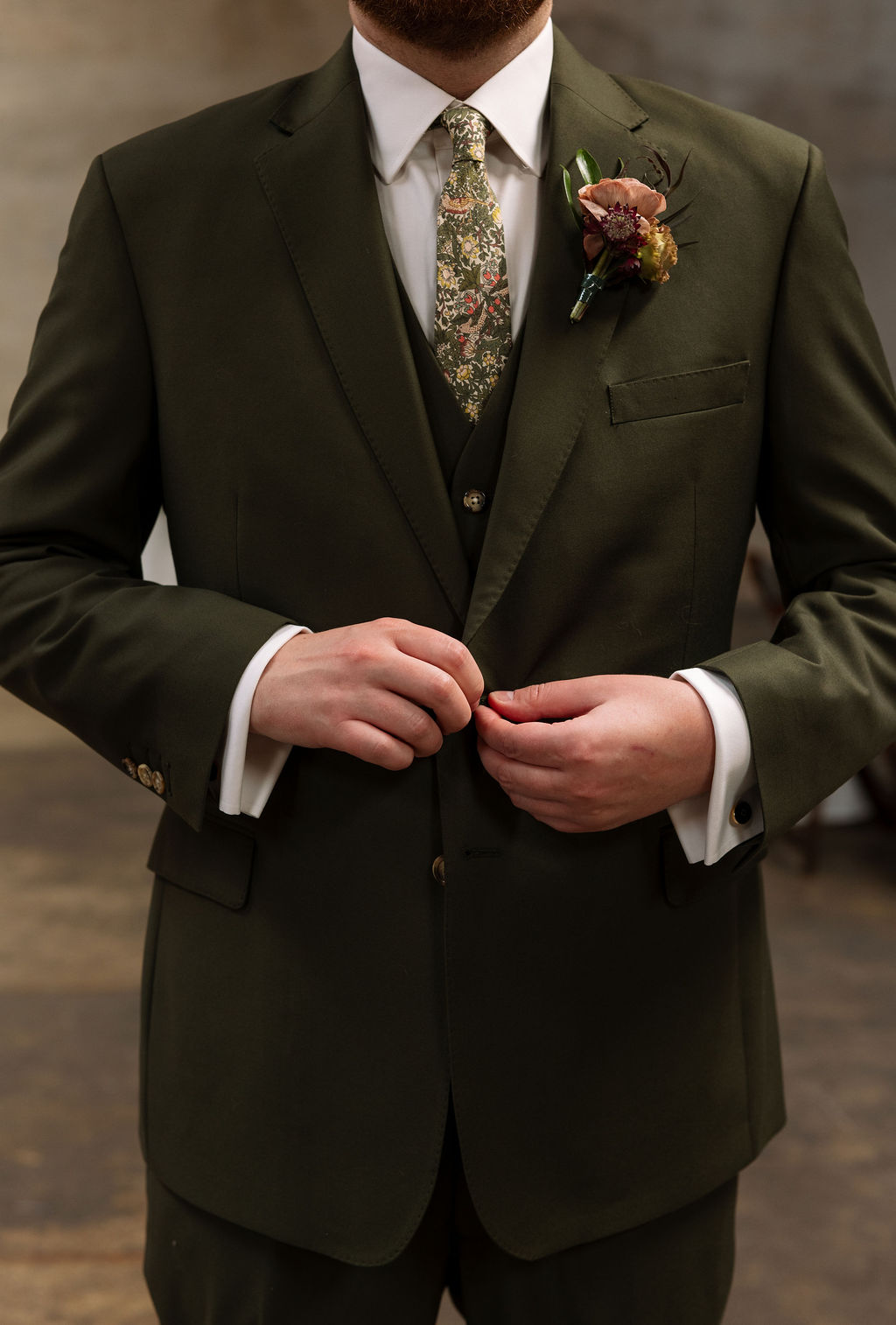 Groom buttoning a deep green suit jacket with floral tie and boutonniere detail