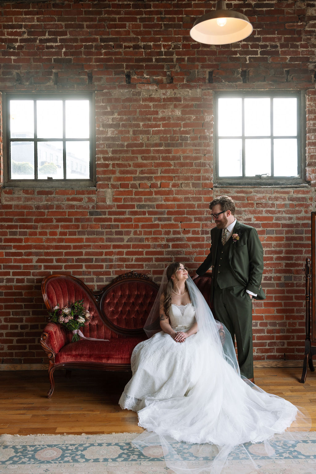 Bride sitting on a vintage couch while groom stands beside her in a brick-walled space, captured by a wedding photographer in Knoxville