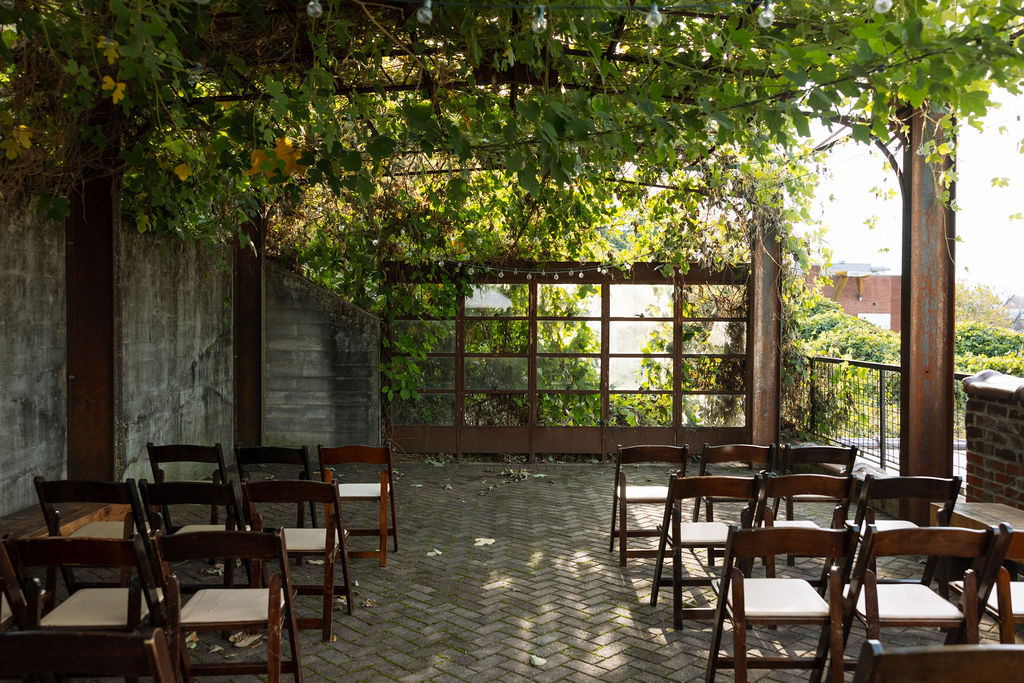 Outdoor ceremony space with wooden chairs under a vine-covered pergola and string lights