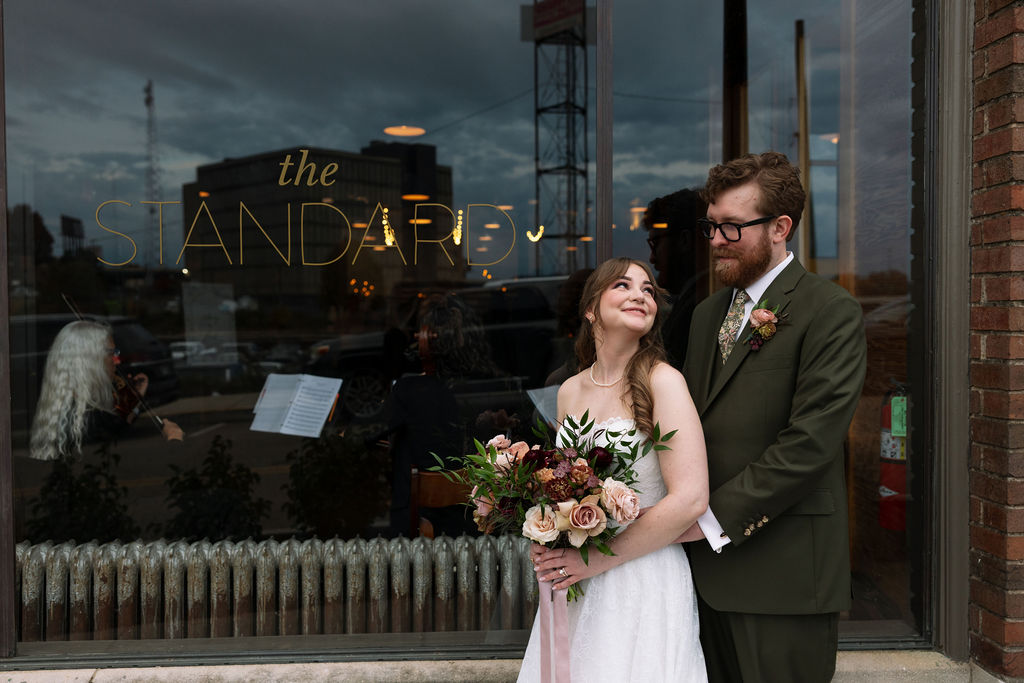 Bride and groom standing outside The Standard venue window with reflections and soft evening light