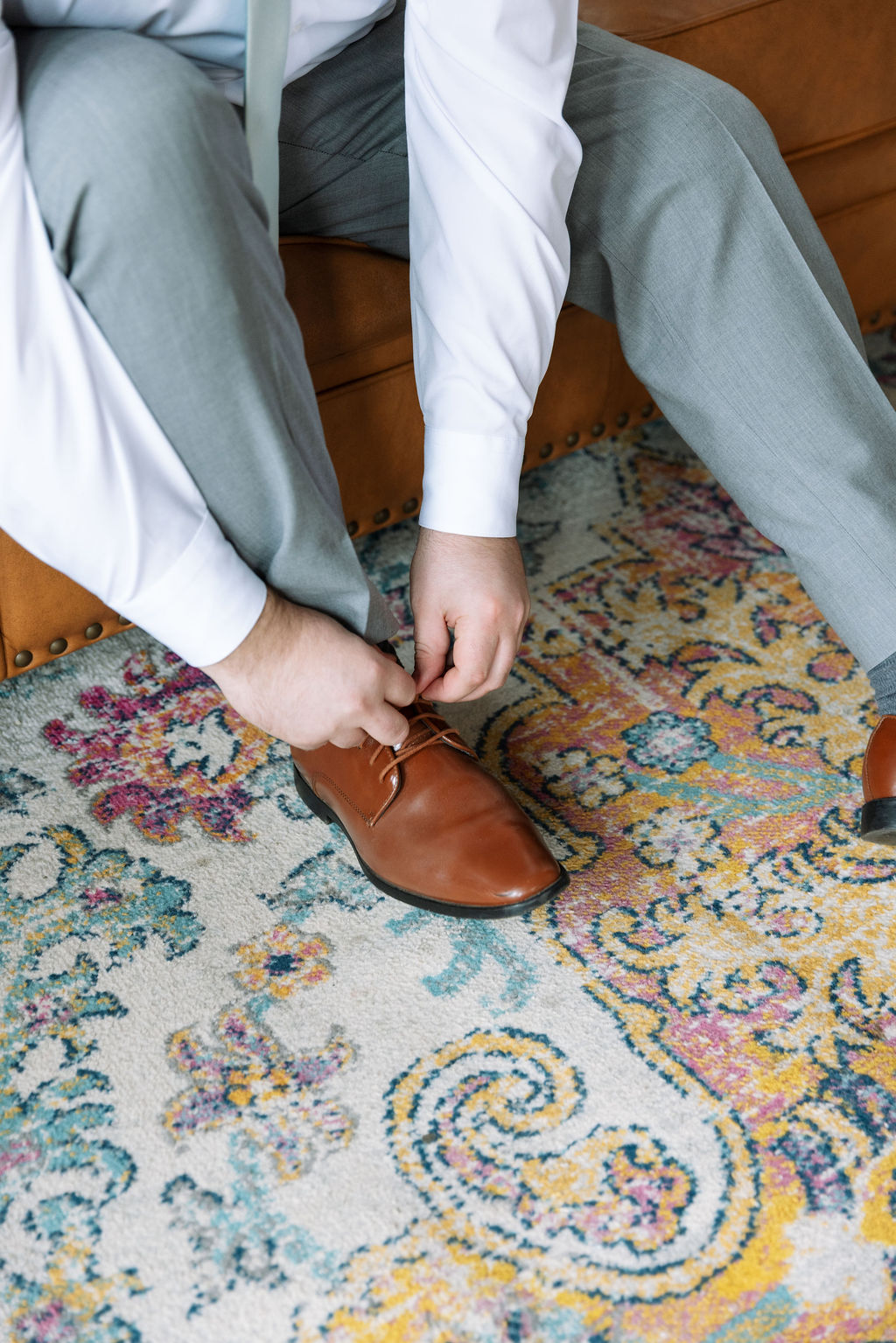 Groom tying brown dress shoes while getting ready on patterned rug before wedding ceremony