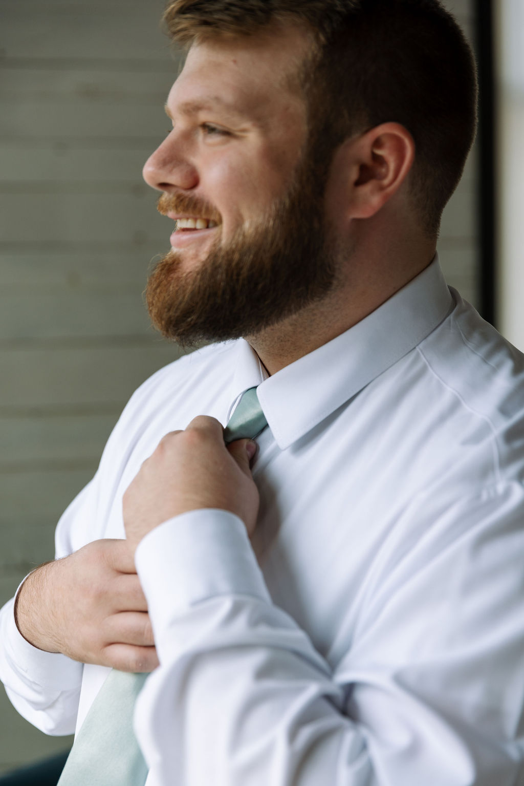 Groom adjusting tie by window during relaxed candid wedding photography getting ready moment