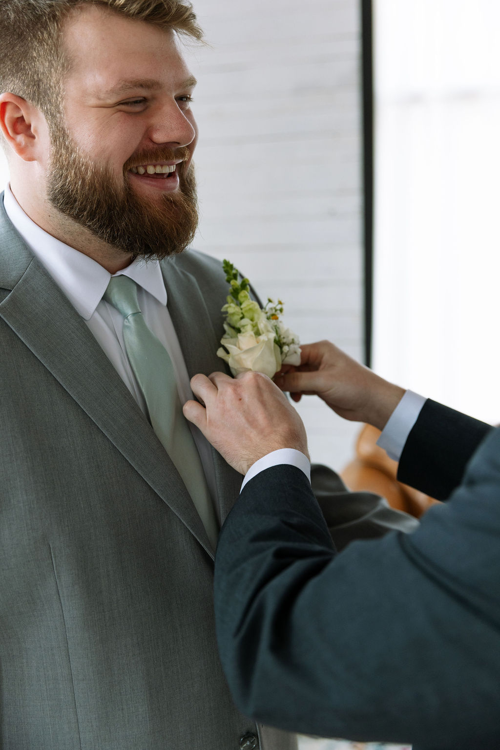 Groomsman helping groom pin boutonniere during candid wedding photography preparation moment