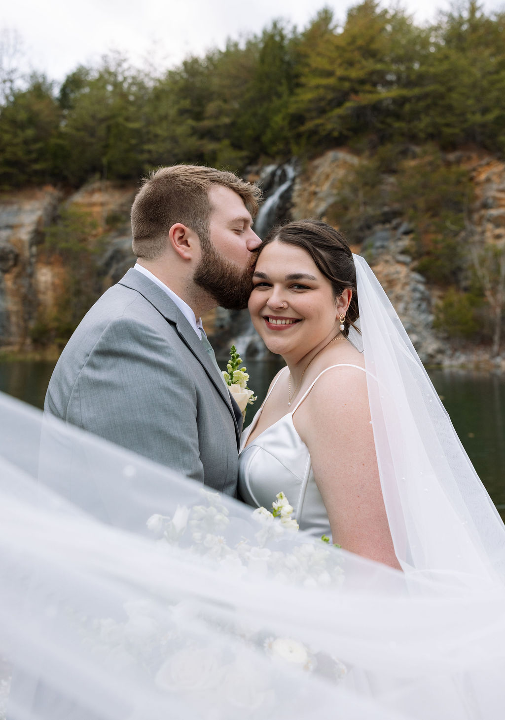 Groom kissing bride’s forehead during waterfall portrait captured with candid wedding photography style