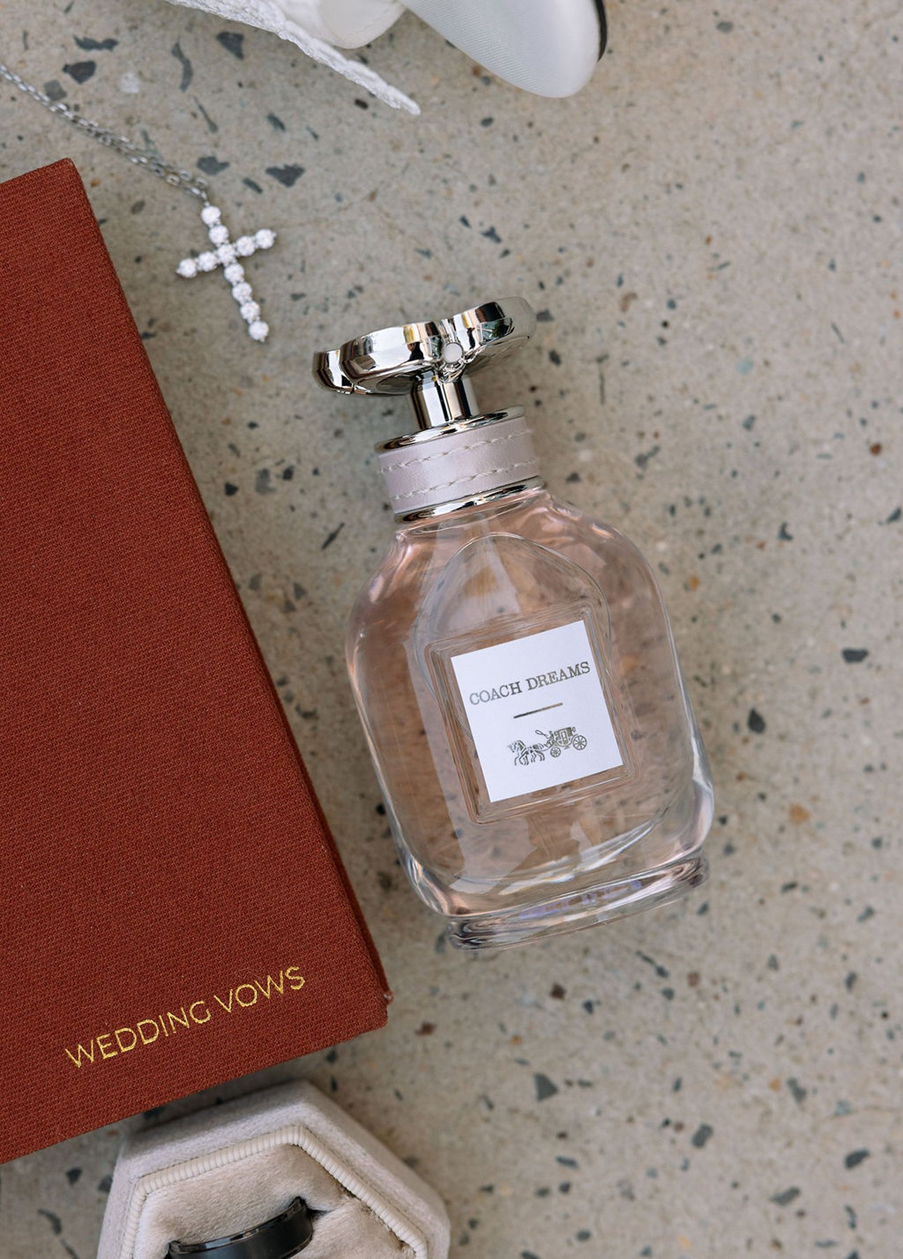 Wedding detail flat lay with vows book, perfume, cross necklace, and ring