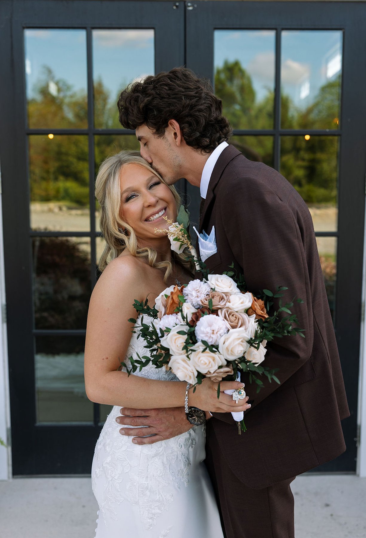 Bride smiling as groom kisses her forehead outside modern venue doors