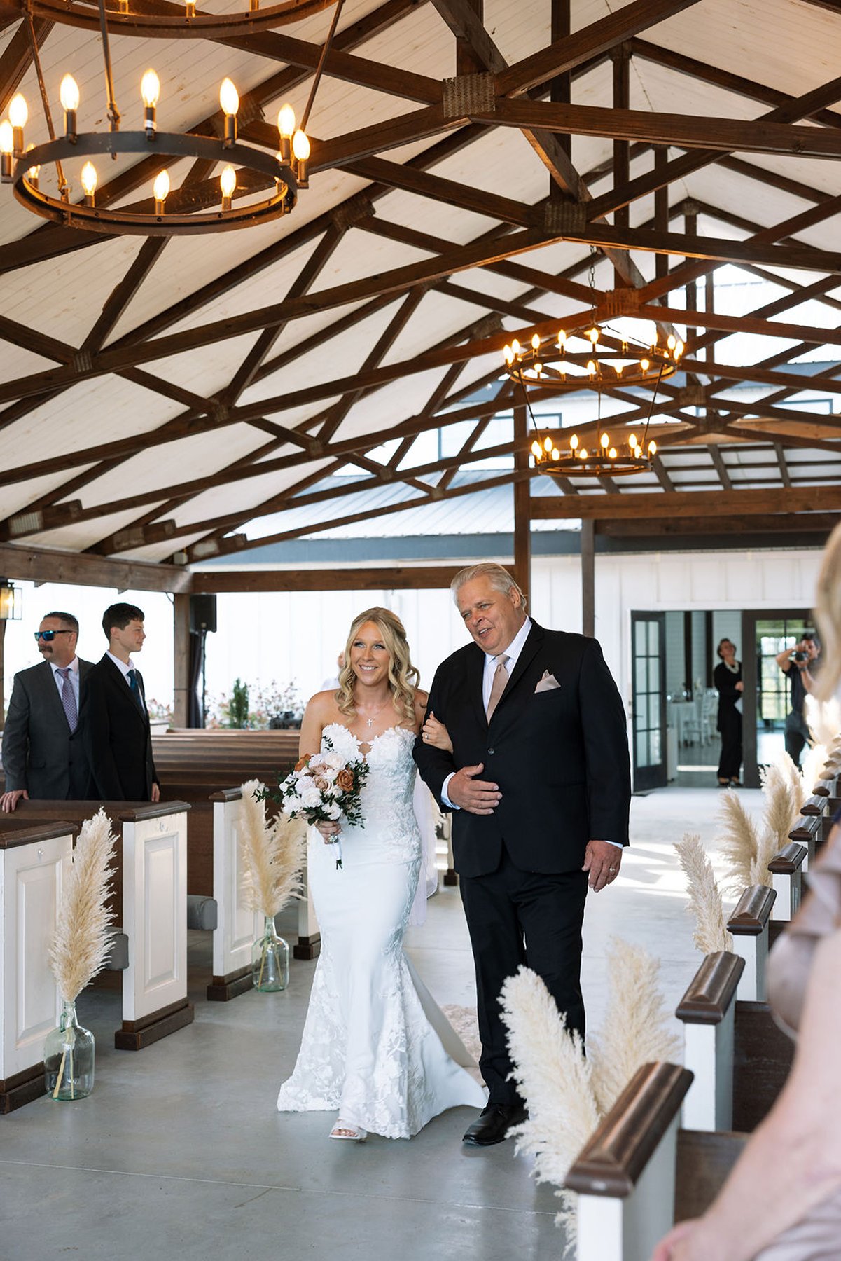 Bride walking down aisle with father inside open-air chapel with wooden beams