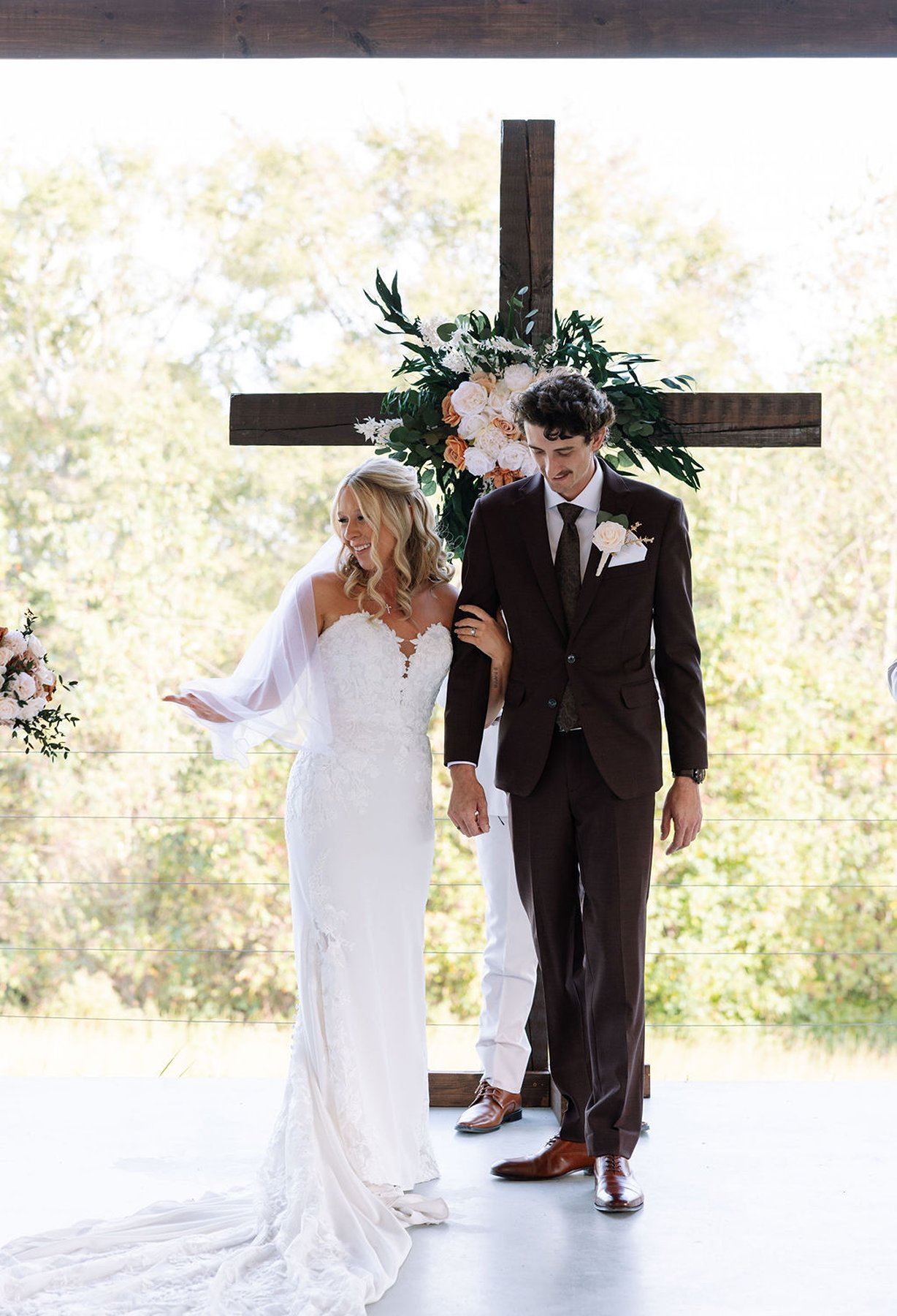 Bride and groom walking together after ceremony beneath wooden cross with florals