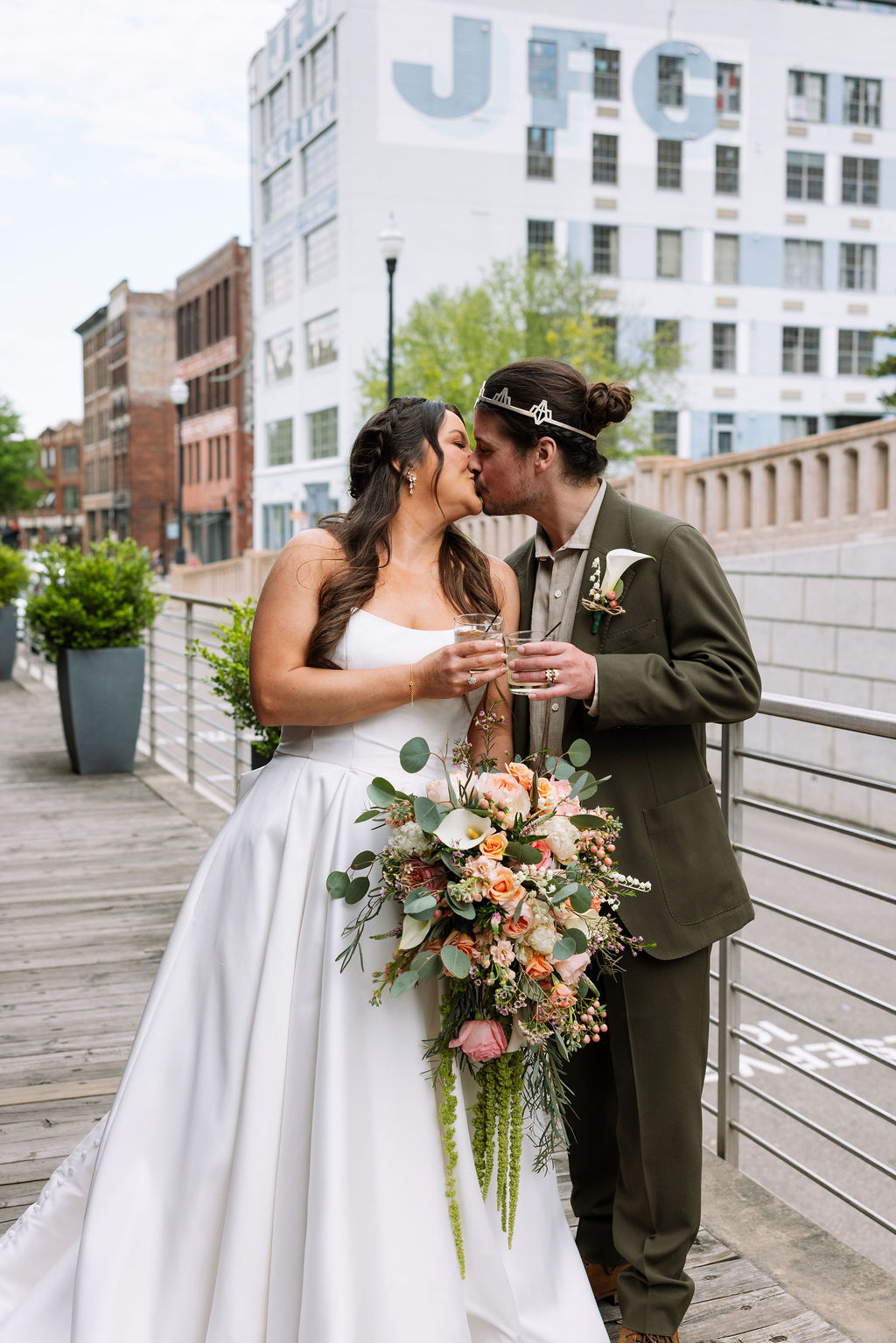 Bride and groom kissing on a downtown Knoxville walkway with city buildings in the background, holding drinks and a colorful bouquet