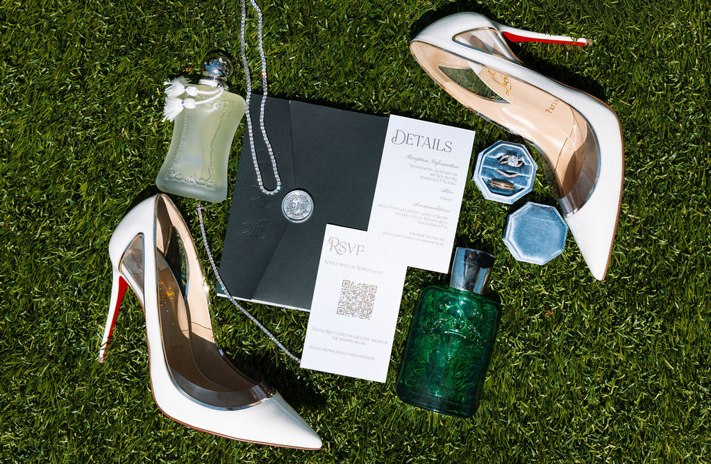 Flat lay of wedding details including invitation suite, shoes, perfume, and rings styled on grass