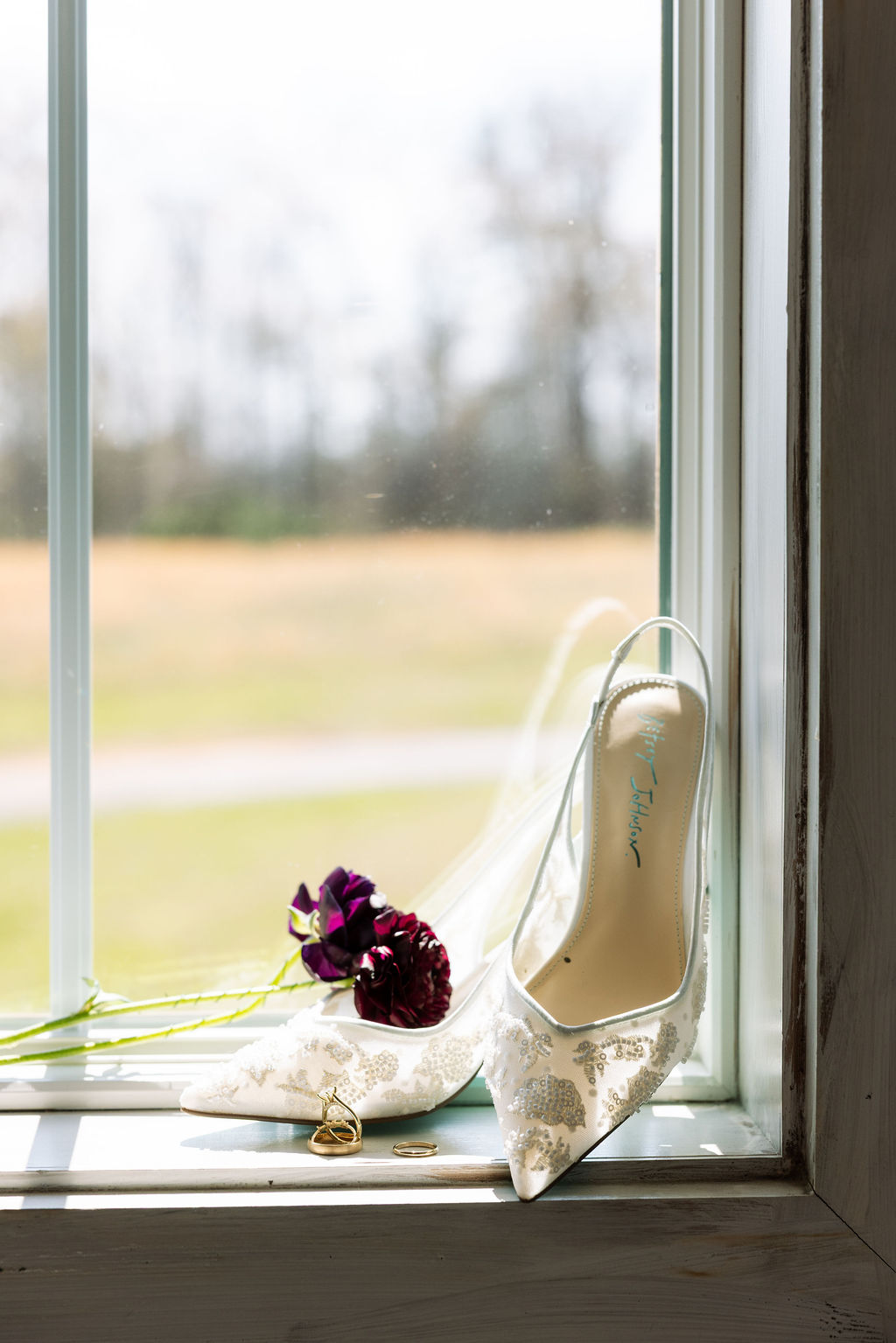 Bridal shoes, rings, and florals arranged by a window with natural light at Howe Farms wedding venue