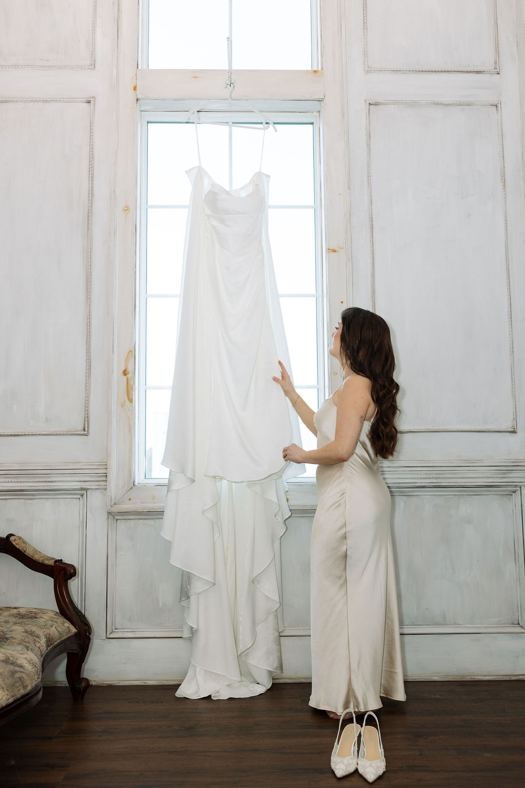 Bride standing by a window reaching for her hanging wedding dress, getting ready at Howe Farms wedding venue
