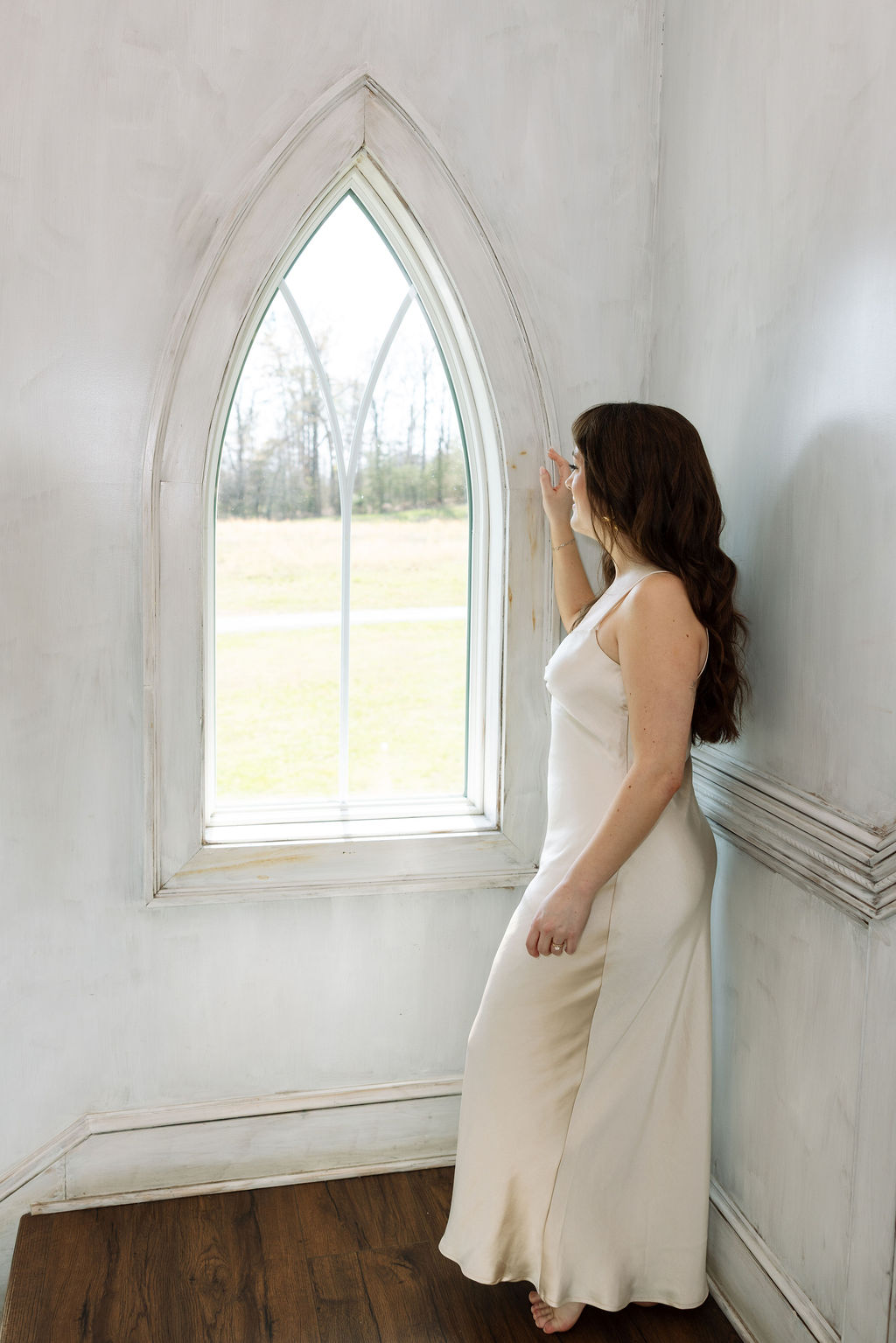 Bride in a silk dress standing by a tall arched window, soft light filling the room