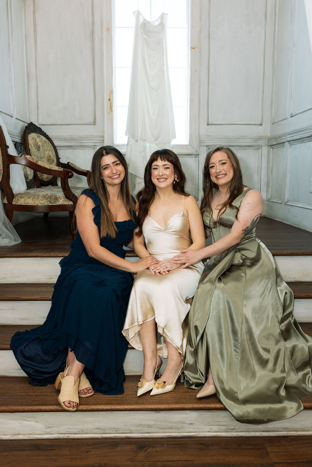 Bride sitting with her bridesmaids on wooden steps beneath her hanging dress at Howe Farms wedding venue