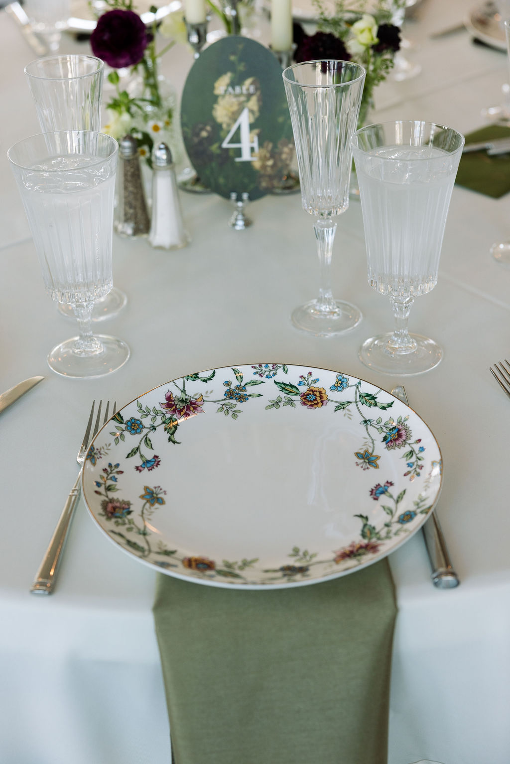 Reception table place setting with floral china, glassware, and soft green linens